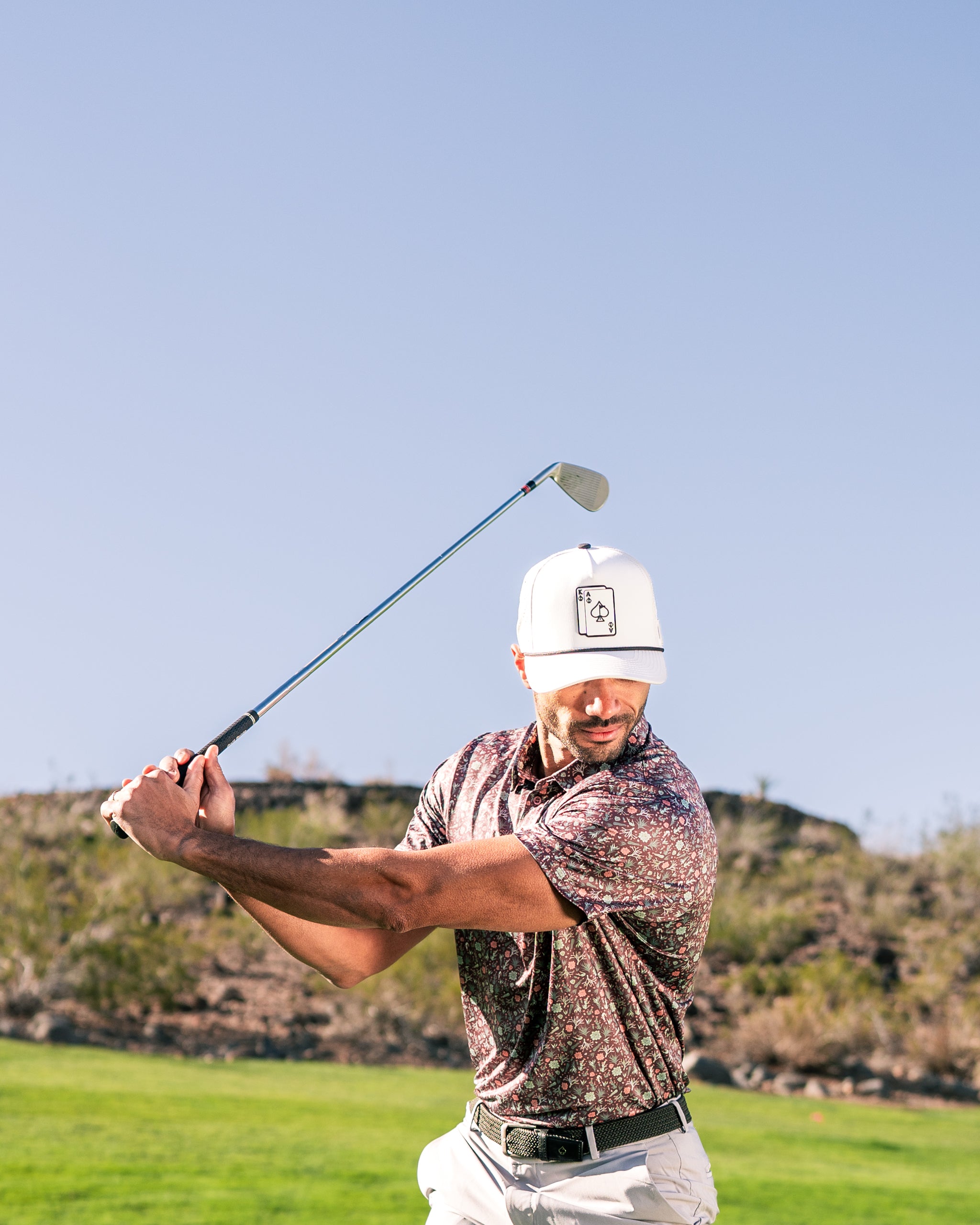 A golfer swings a club on a sunny course, wearing a floral-patterned shirt and a white cap, showcasing bold golf style.