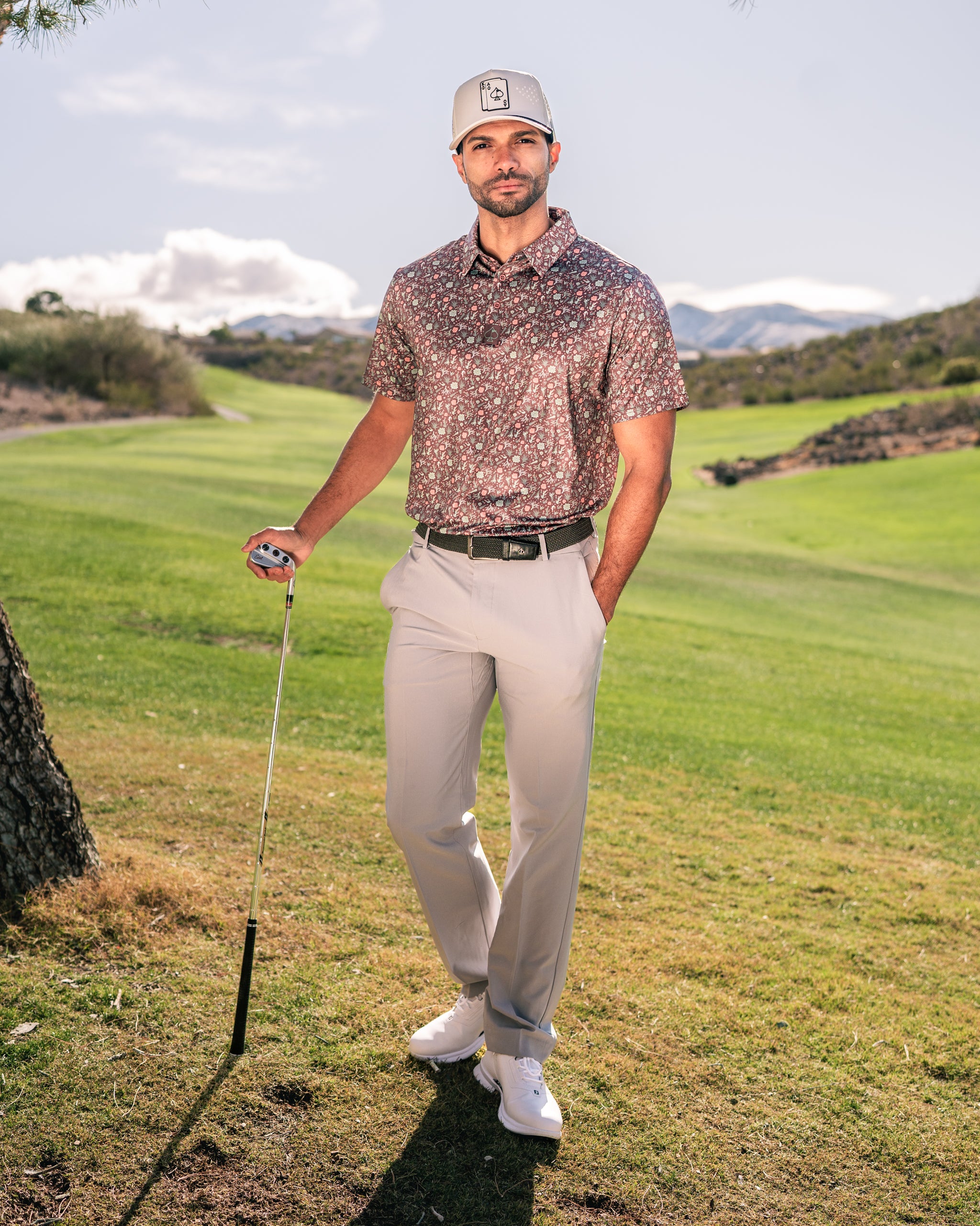 A golfer stands confidently on a lush green course, wearing a Desert Sage floral polo shirt, light pants, and a cap.