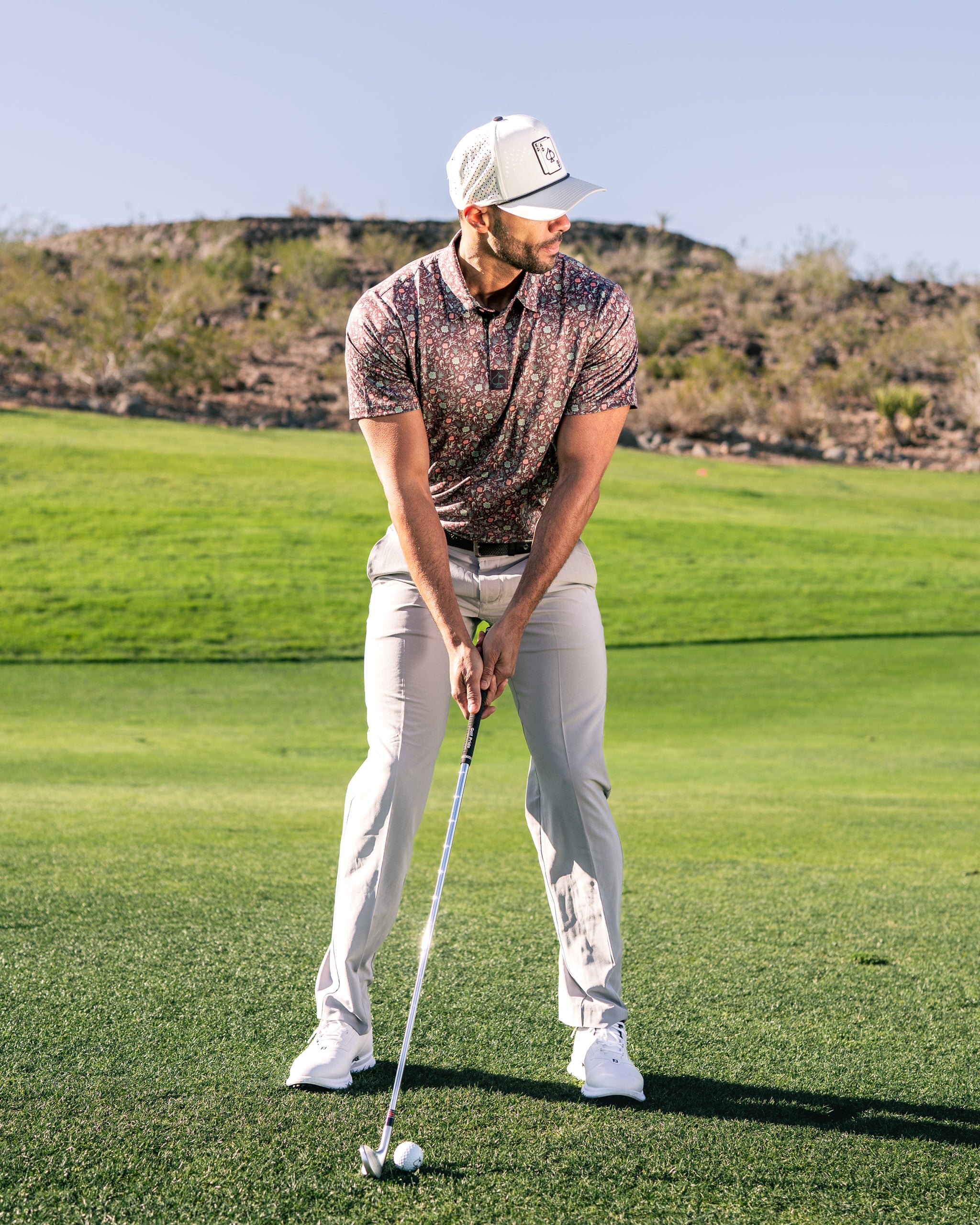 A golfer in a floral-patterned polo shirt and light pants prepares to swing on a sunny golf course.