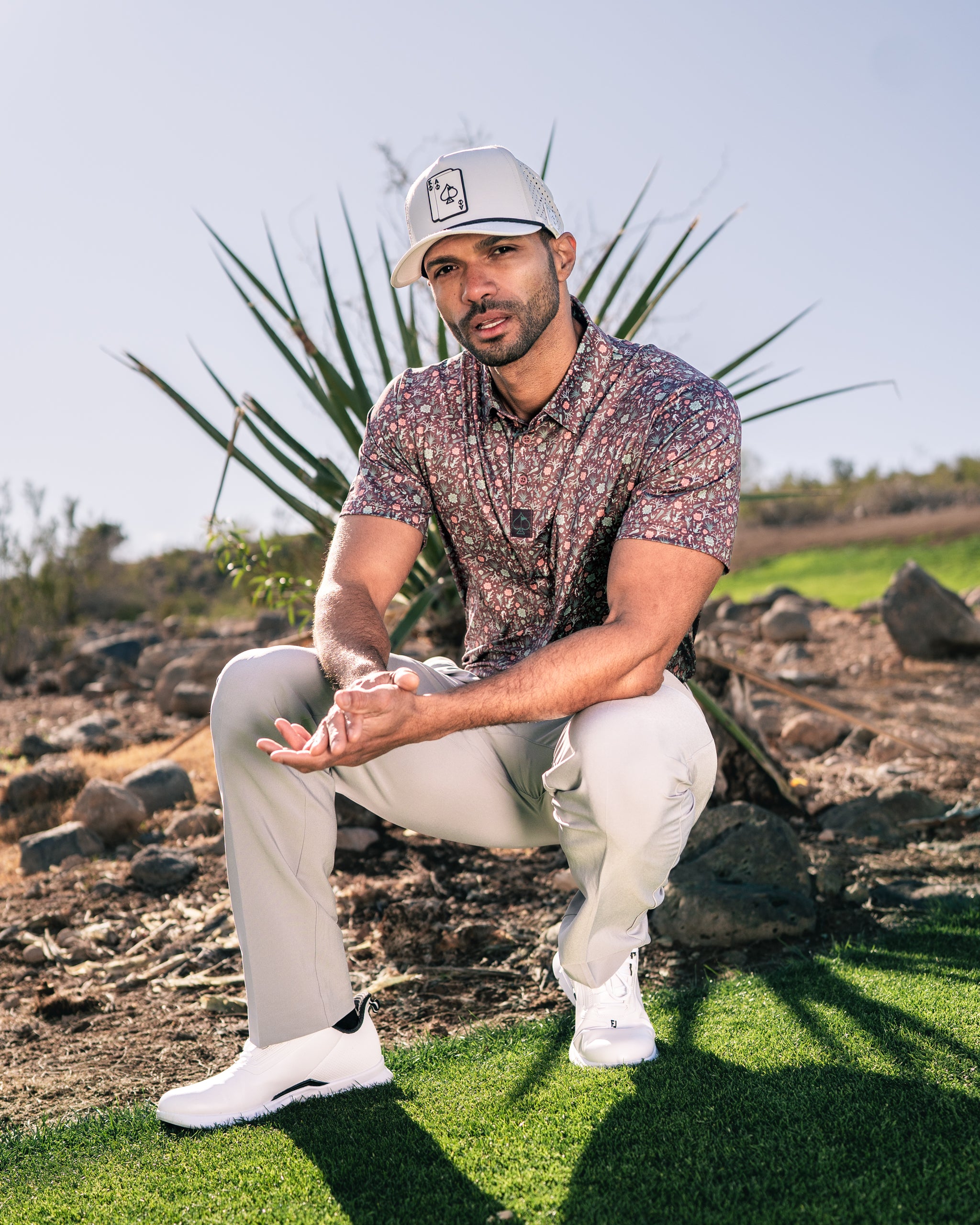 Man in a floral golf shirt and cap kneels on a golf course, showcasing the Desert Sage apparel against a desert backdrop.