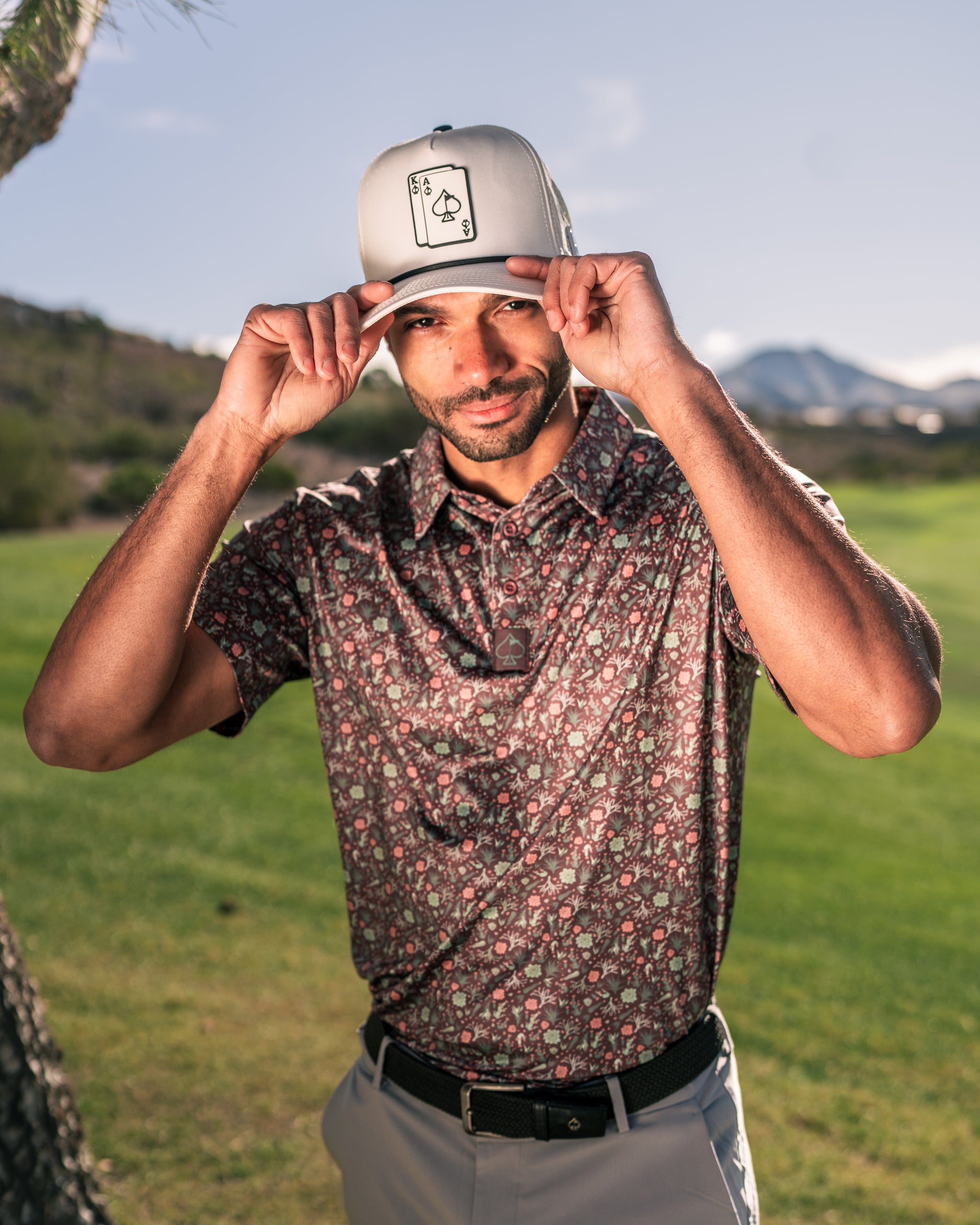 Man wearing a floral golf shirt and a gray cap with a spade logo, posing on a golf course with mountains in the background.