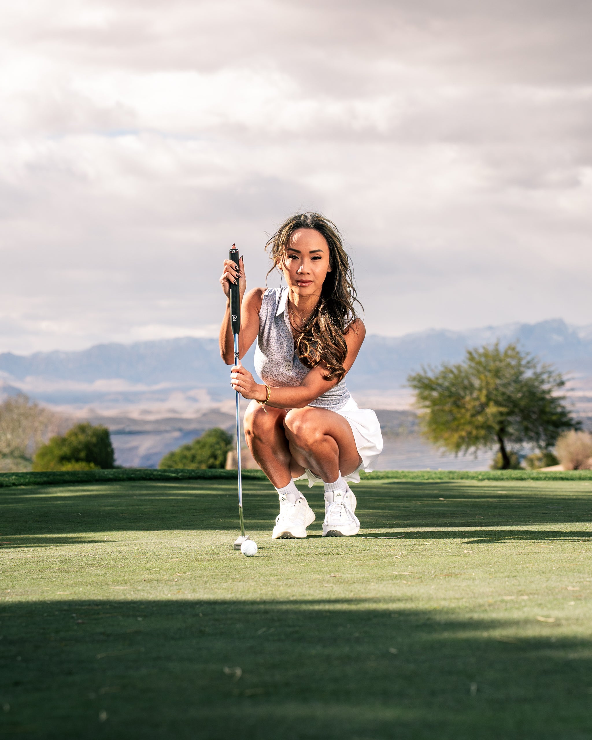 A woman in a stylish golf outfit kneels on the green, preparing to putt, showcasing the Women's Best Friend Light apparel.
