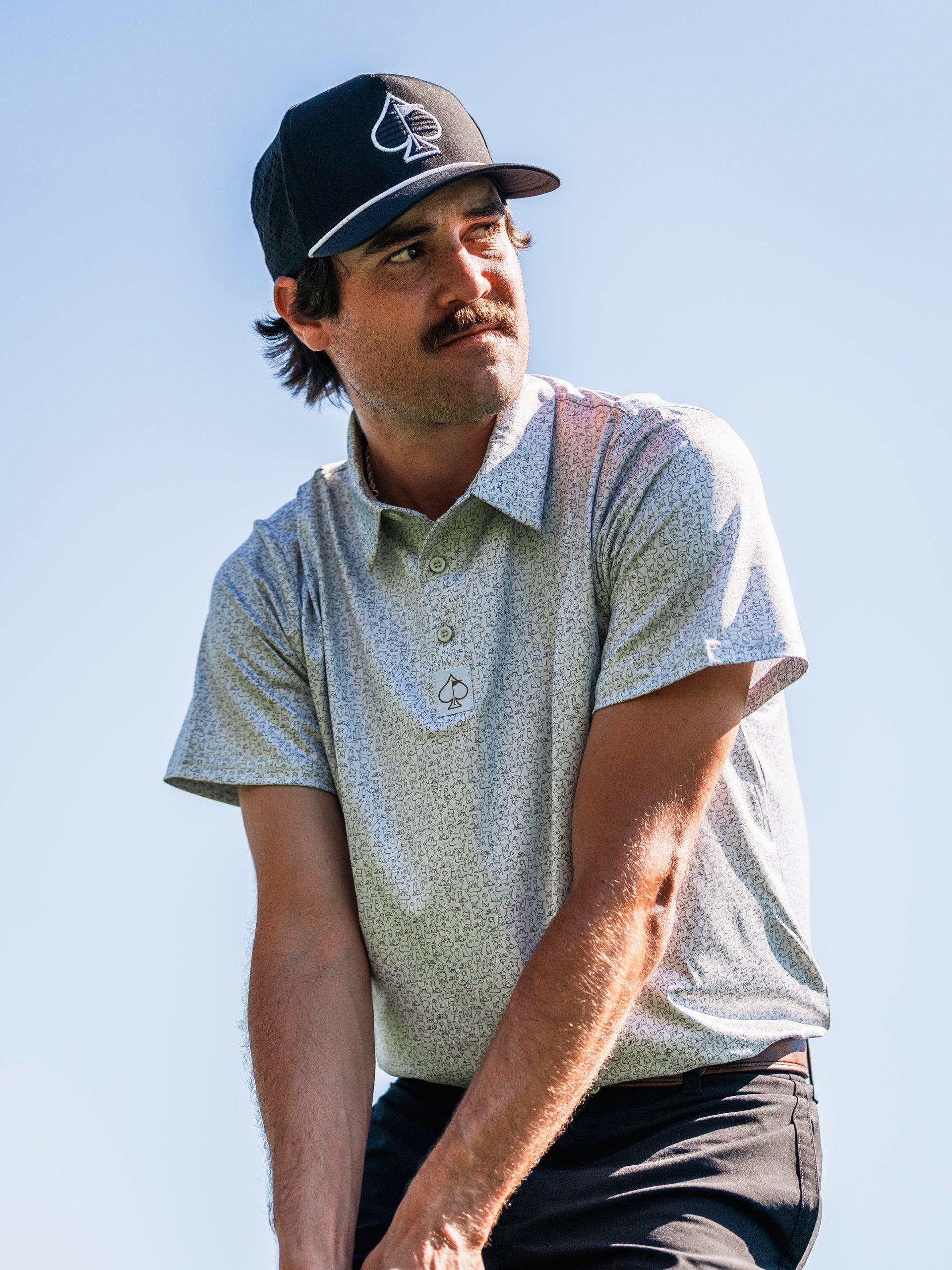 A golfer wearing a light-patterned polo shirt and a black cap prepares to swing on the course, showcasing stylish golf apparel.