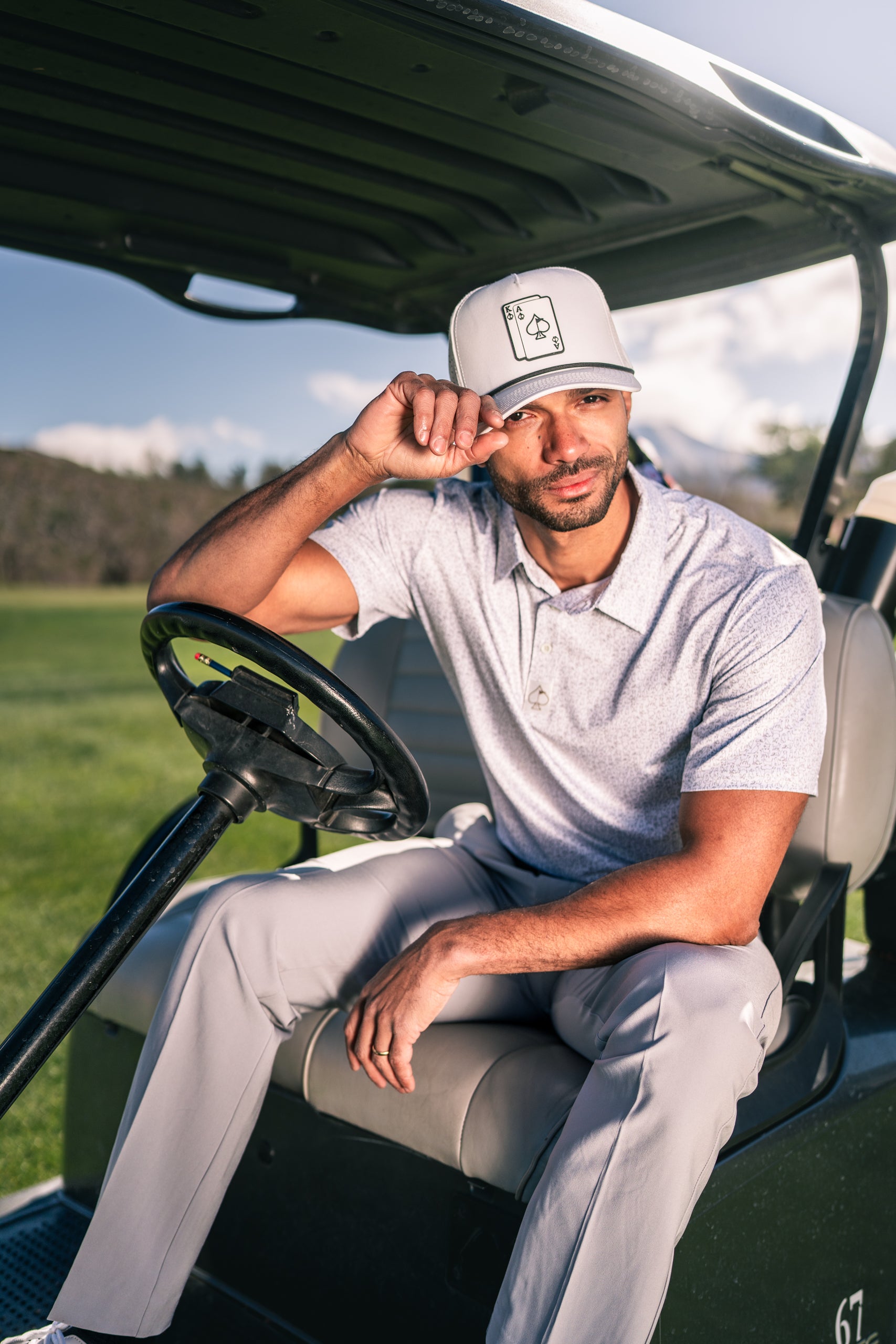 Man sitting in a golf cart, wearing a light gray polo and a cap featuring a dog graphic, enjoying a sunny day on the course.