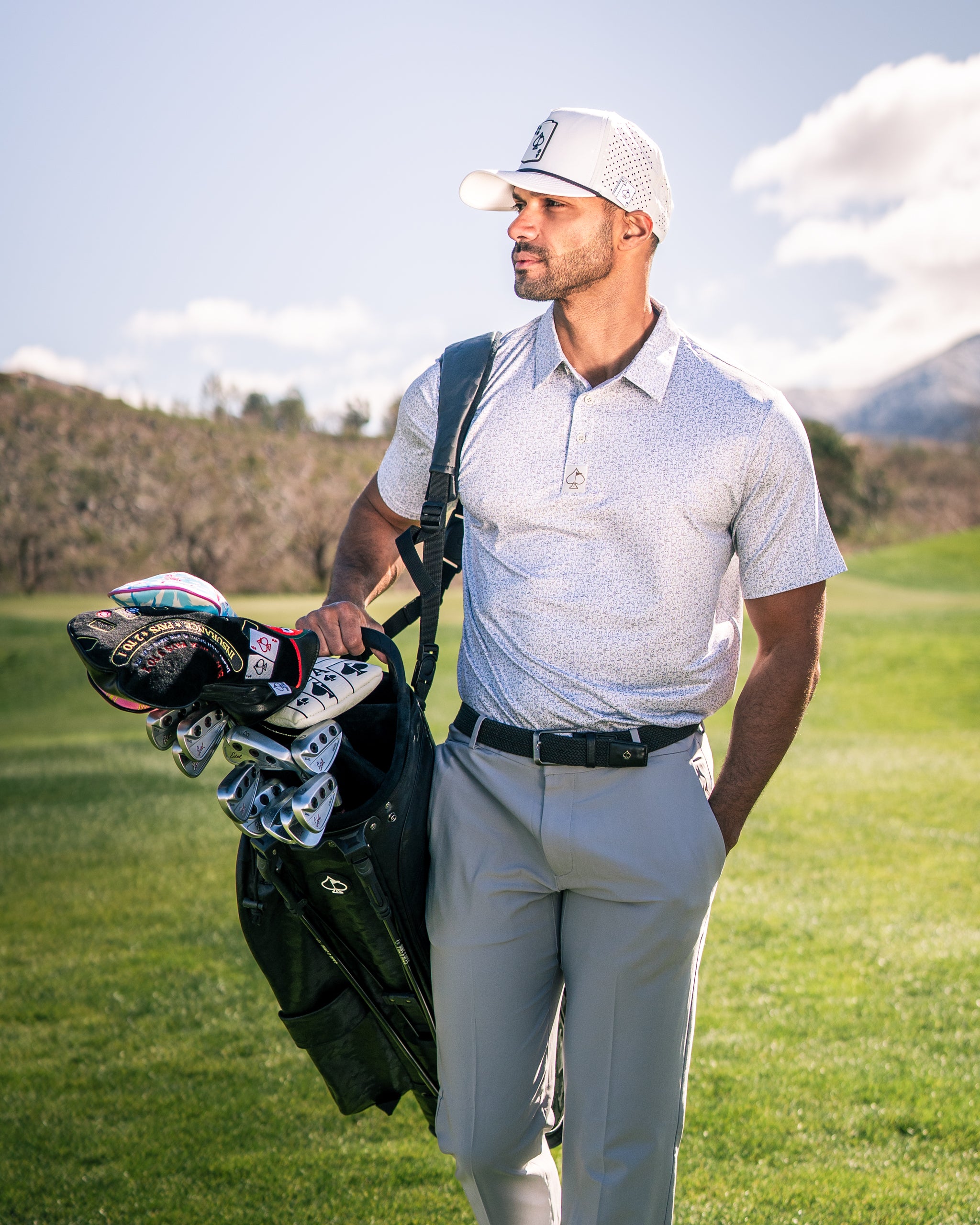 A golfer in a stylish polo shirt and cap carries a golf bag with colorful headcovers on a sunny course.
