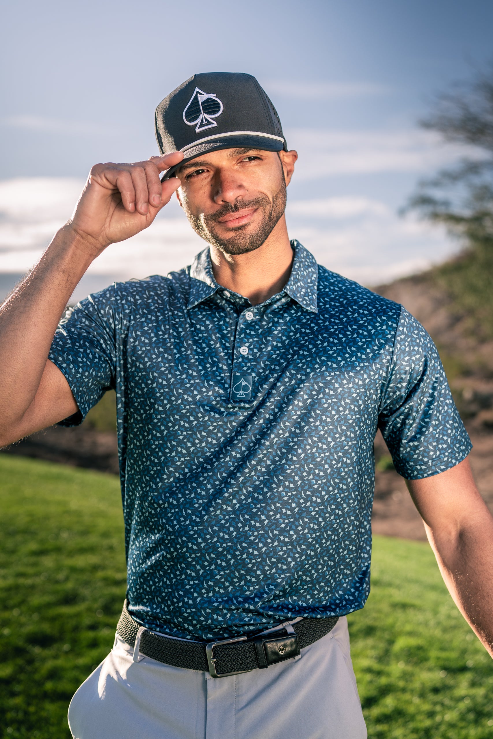Man in a blue patterned golf polo and cap, smiling while adjusting his hat on a golf course.