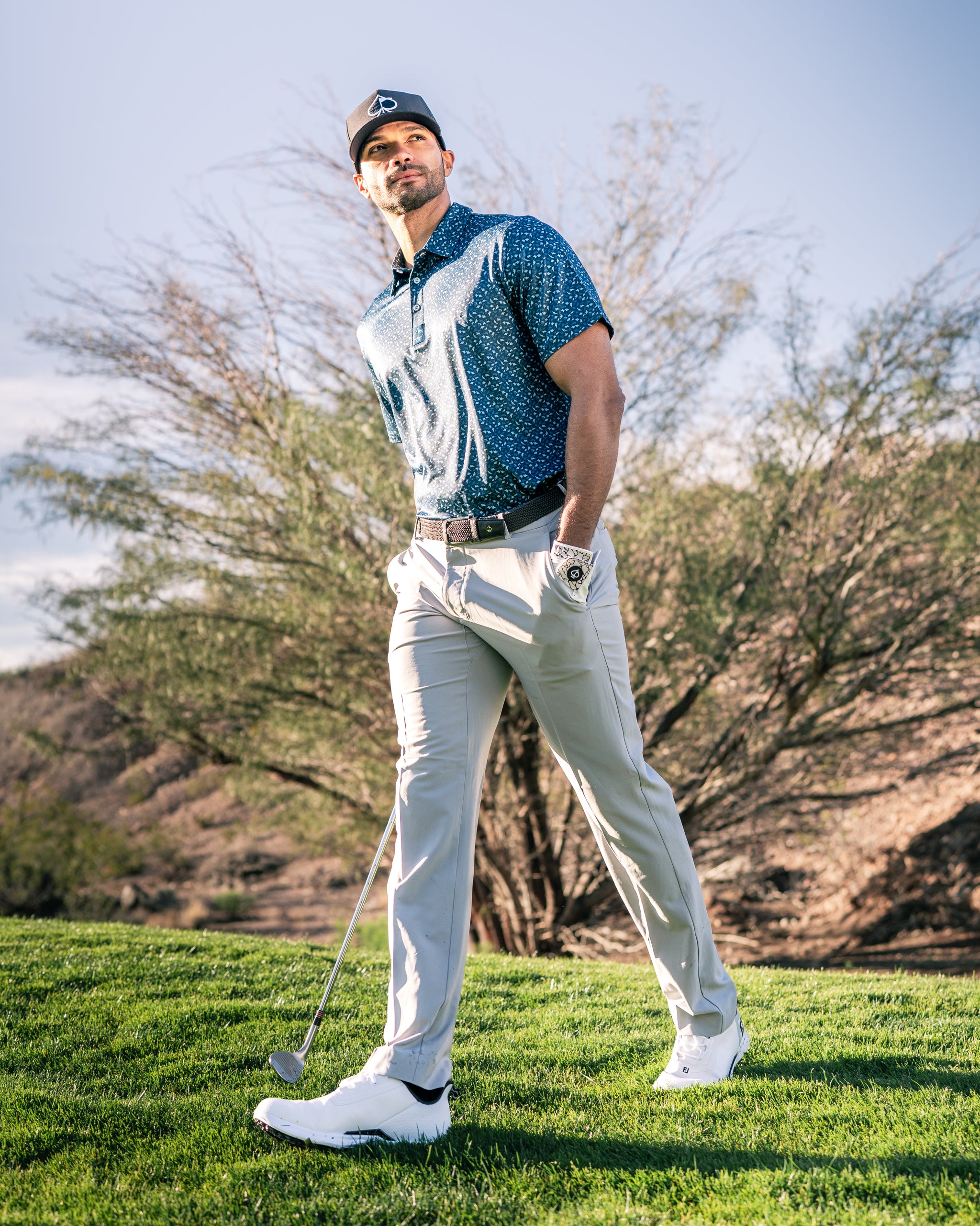 A golfer wearing a patterned blue polo and beige pants walks confidently on the course, holding a golf club.
