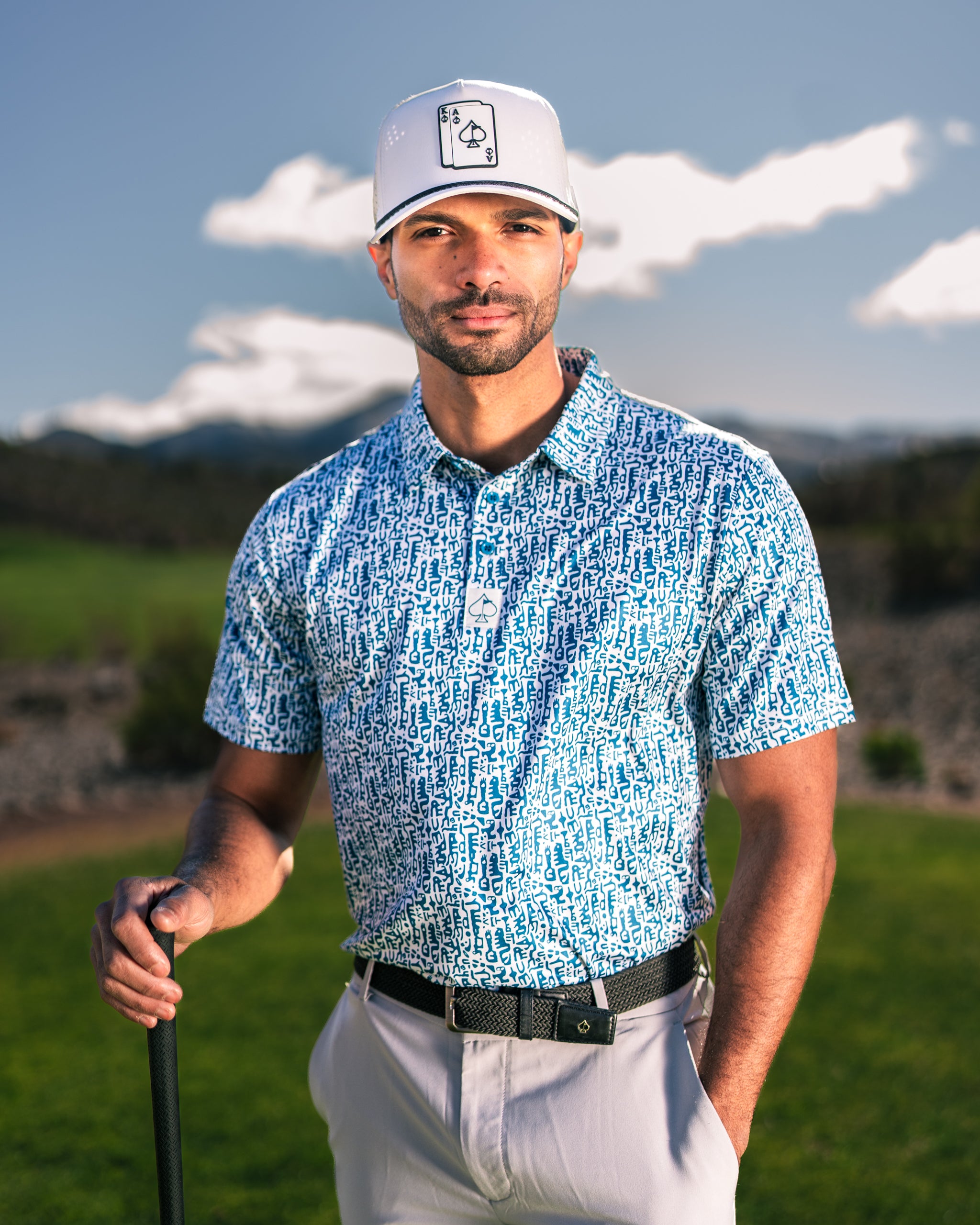 A man stands on a golf course wearing a blue and white patterned polo shirt and a gray cap, holding a golf club.