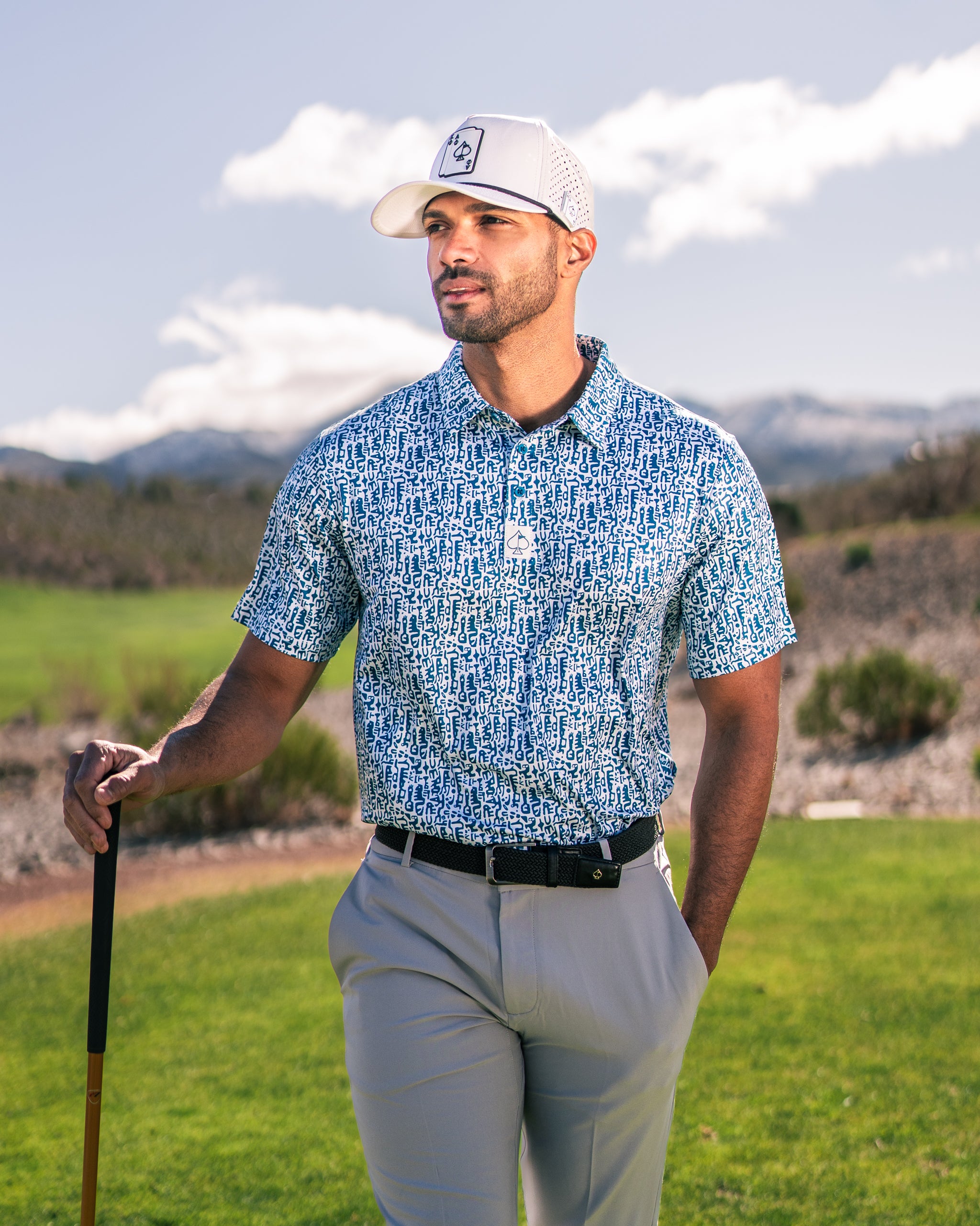 Stylish golfer wearing a blue Maze Craze polo shirt and cap, posing on a sunny golf course with mountains in the background.