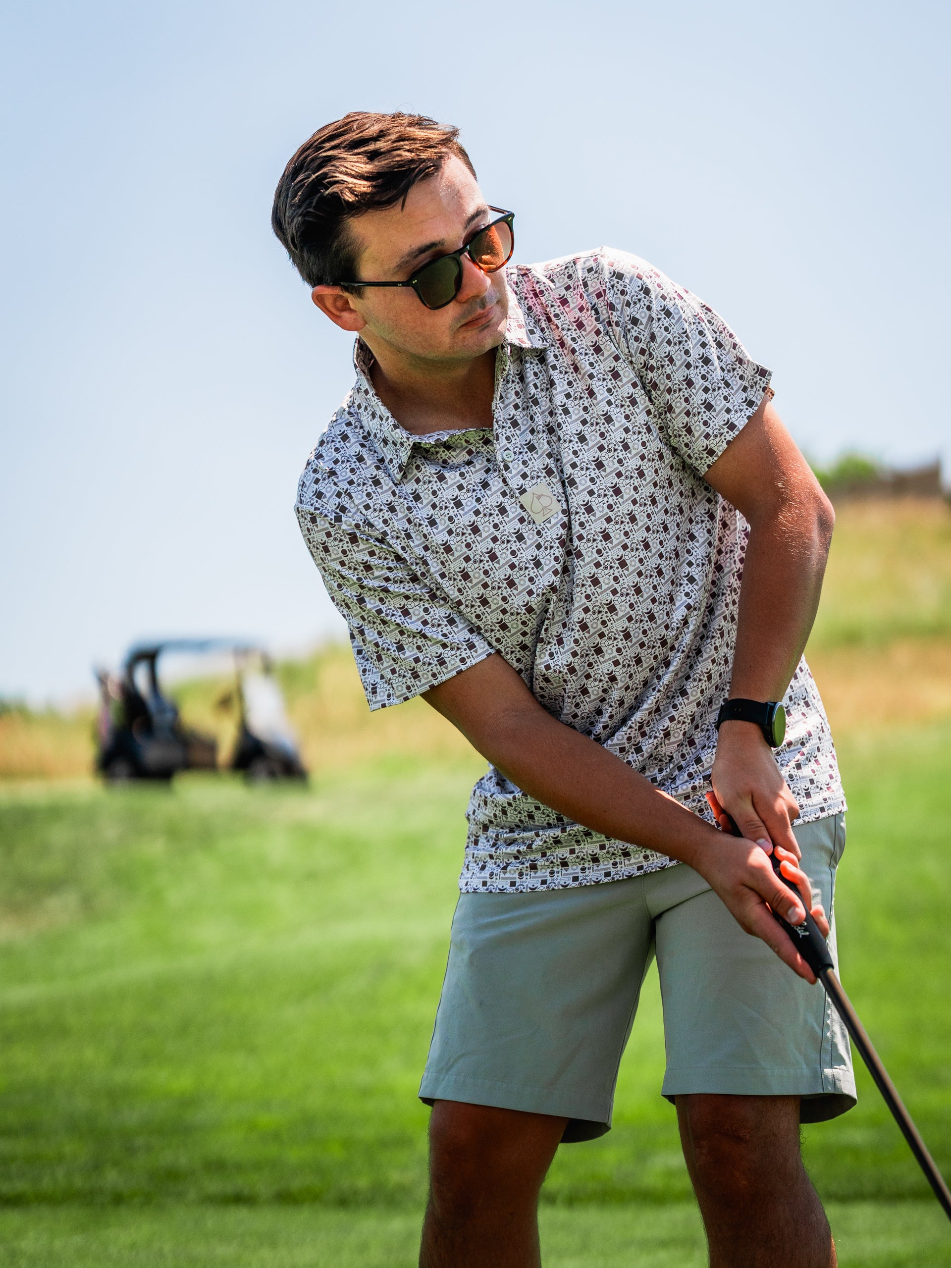 A golfer wearing a retro-patterned short-sleeve shirt and sunglasses prepares to swing on a sunny course.