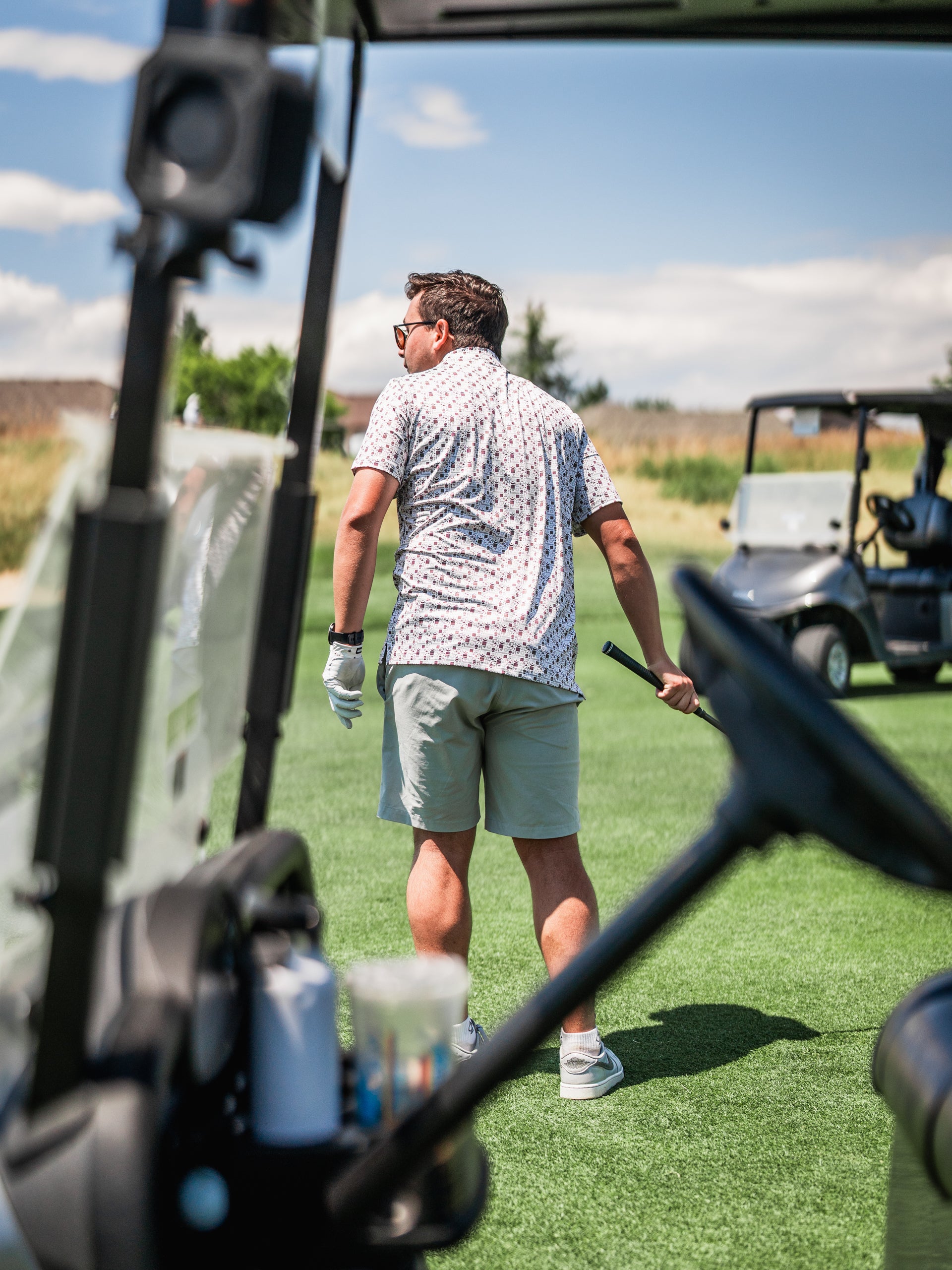 A golfer wearing a patterned shirt and shorts stands on the course, preparing to swing a club near a golf cart.