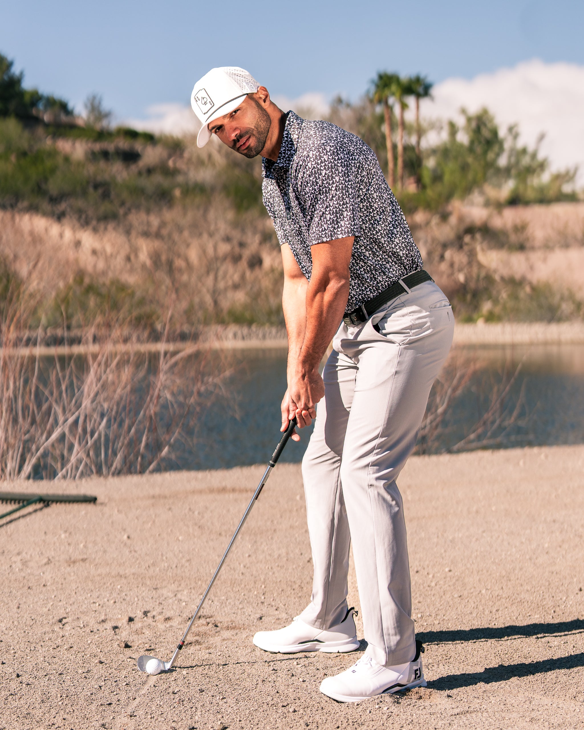 A golfer prepares to swing on a sandy course, wearing a stylish floral polo and light pants, showcasing bold golf fashion.
