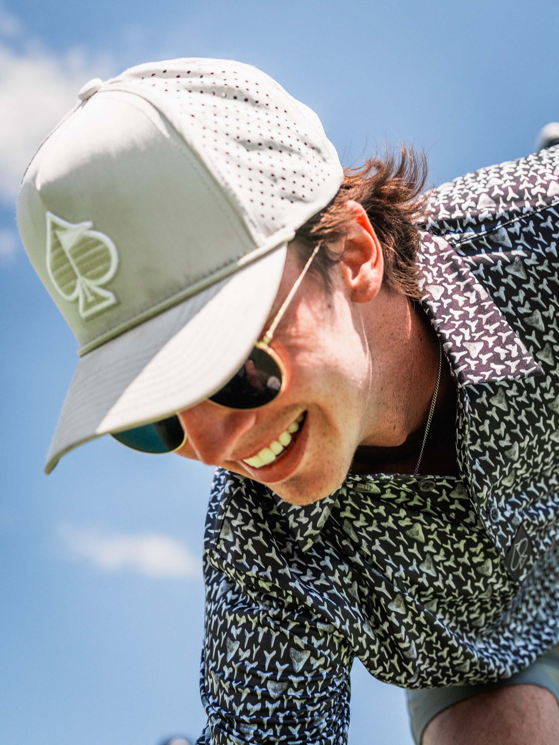 A golfer wearing a Shark Tooth cap and stylish shirt smiles while focusing on the green under a clear blue sky.