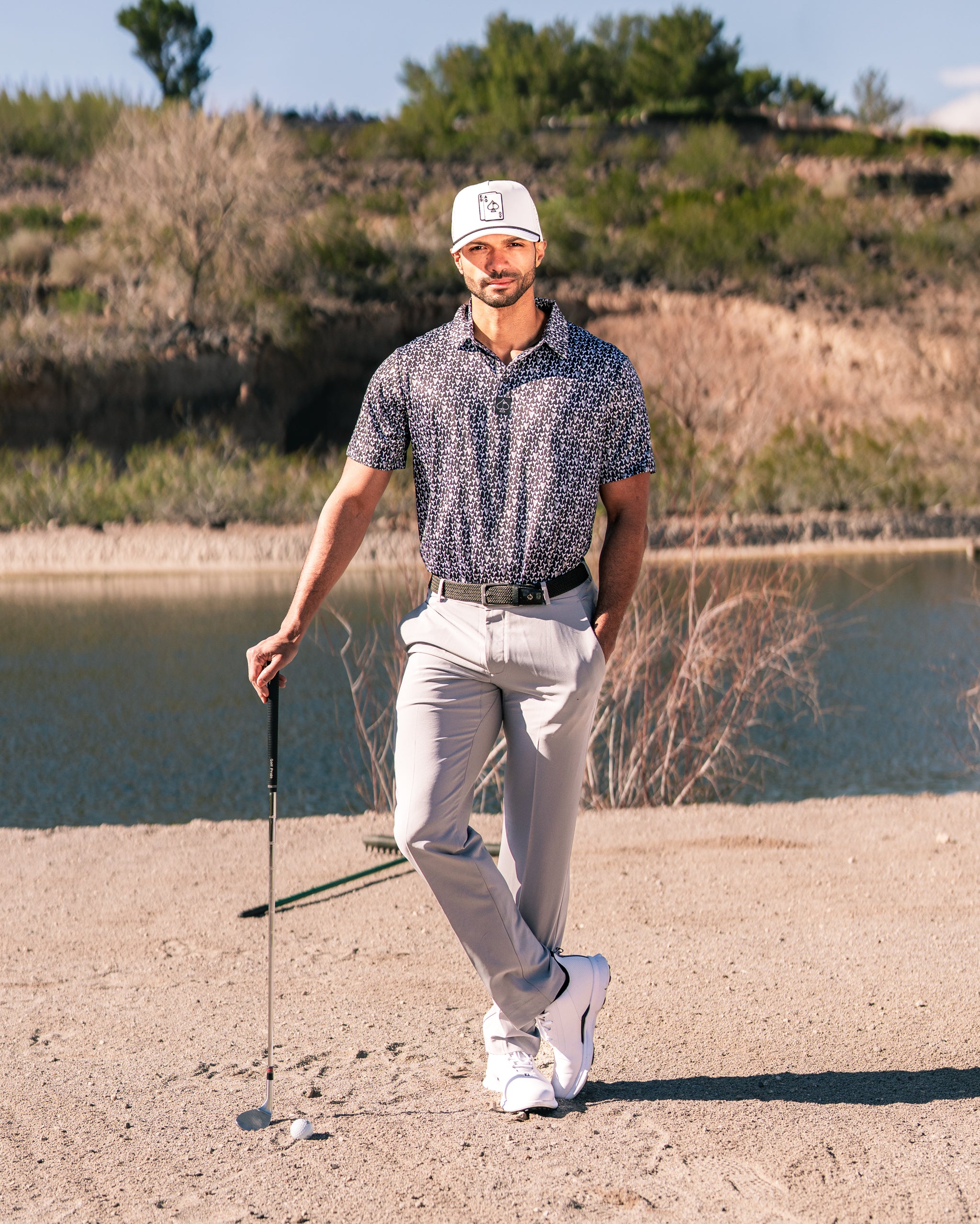 Stylish golfer wearing a patterned shirt and light pants, posing with a golf club near a sandy area by a lake.