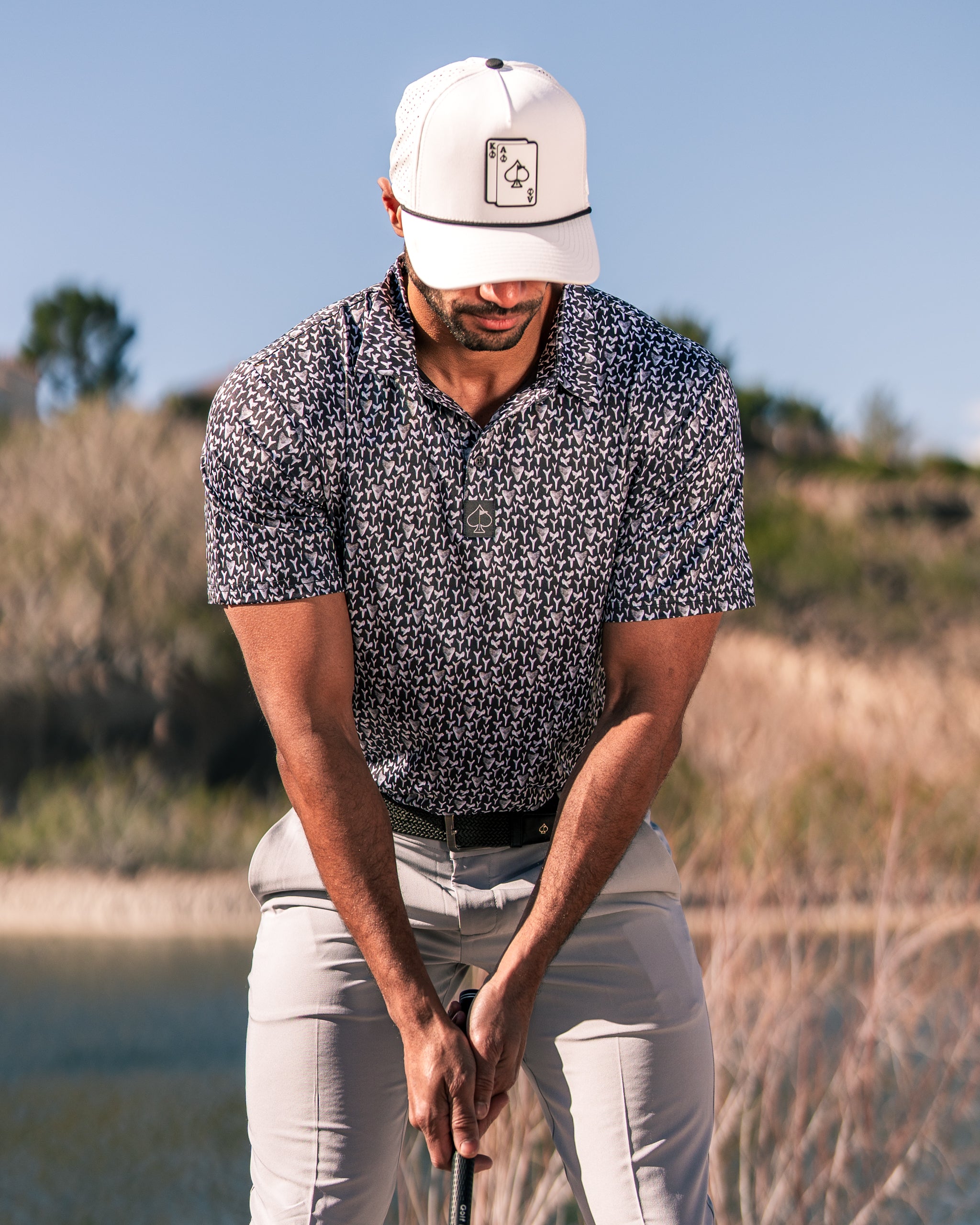 Stylish golfer wearing a patterned shirt and cap, preparing to swing a golf club near a scenic water backdrop.