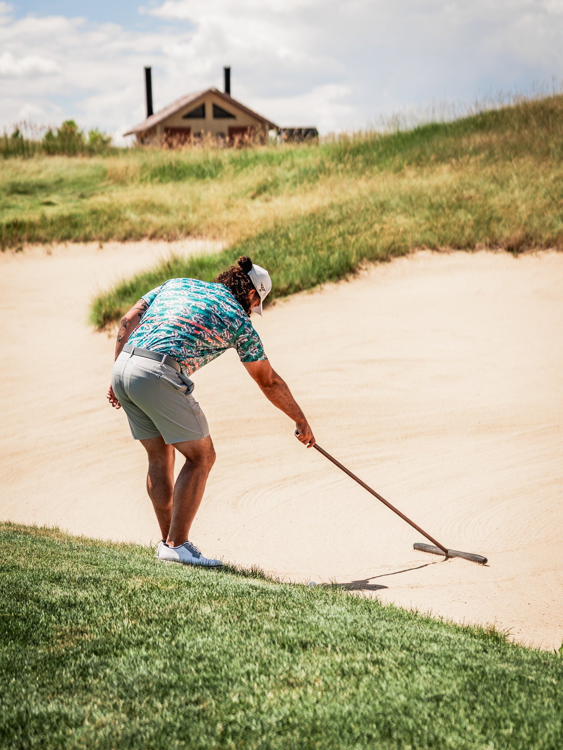 Man in colorful Trout Splatter golf shirt and shorts raking a sand trap on a golf course under a cloudy sky.