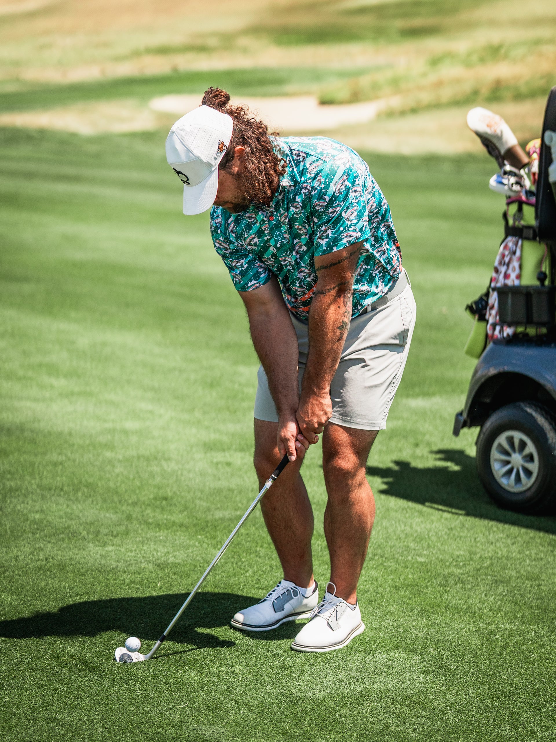 A golfer in a colorful trout-patterned shirt and shorts prepares to chip a ball on the green, showcasing bold golf style.