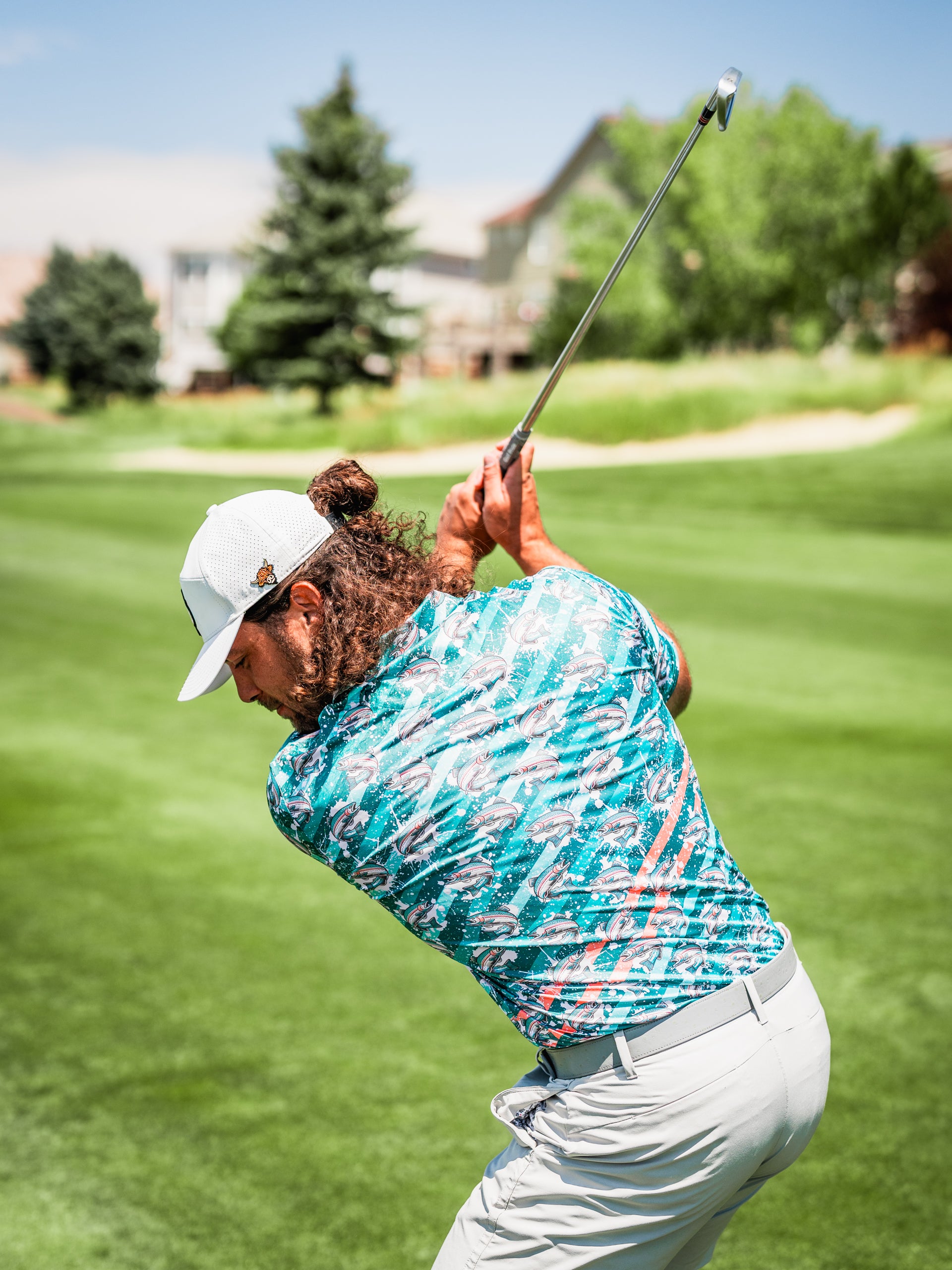 A golfer swings on the course, wearing a vibrant Trout Splatter shirt with a colorful fish pattern, showcasing bold style.