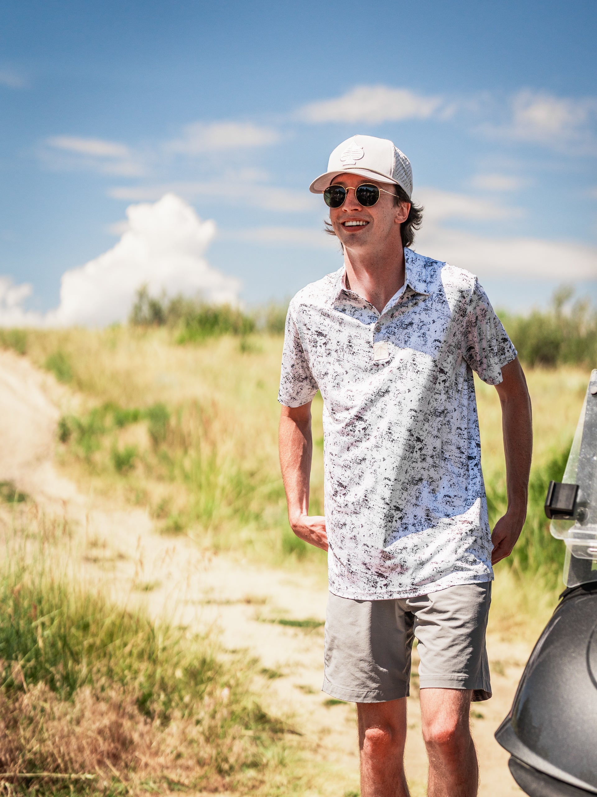 A man wearing a white floral golf polo and gray shorts stands outdoors, smiling against a sunny backdrop.