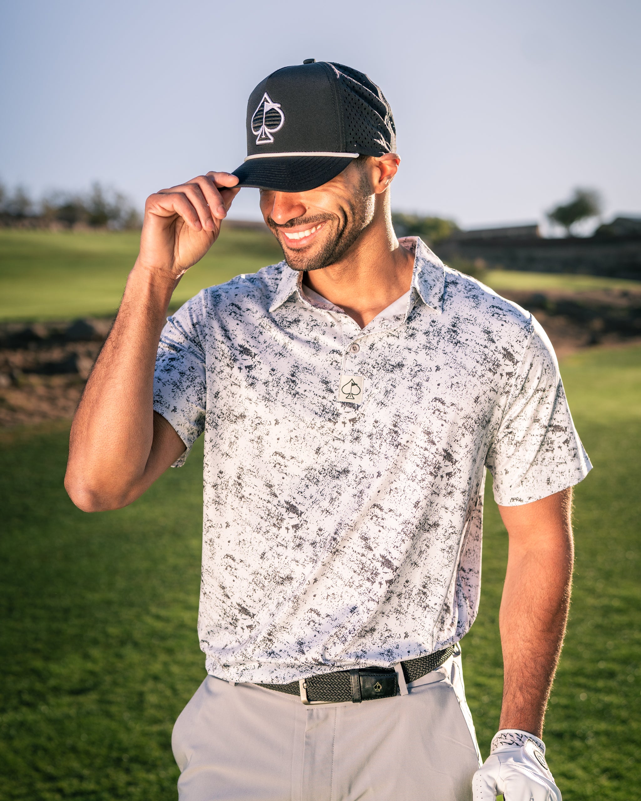 A golfer wearing a stylish black cap and a white patterned polo shirt smiles while adjusting his hat on the course.