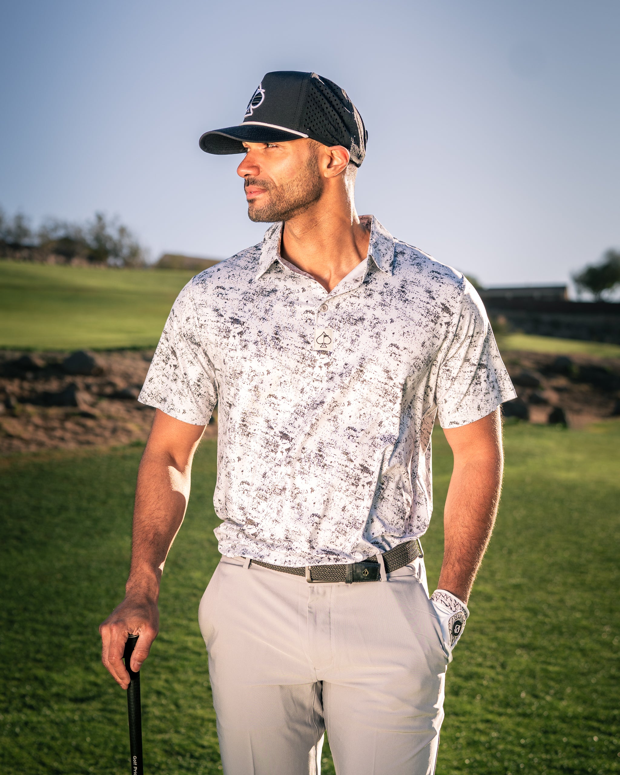 A golfer stands confidently on the course wearing a stylish white and gray patterned polo shirt, paired with light-colored pants and a cap.