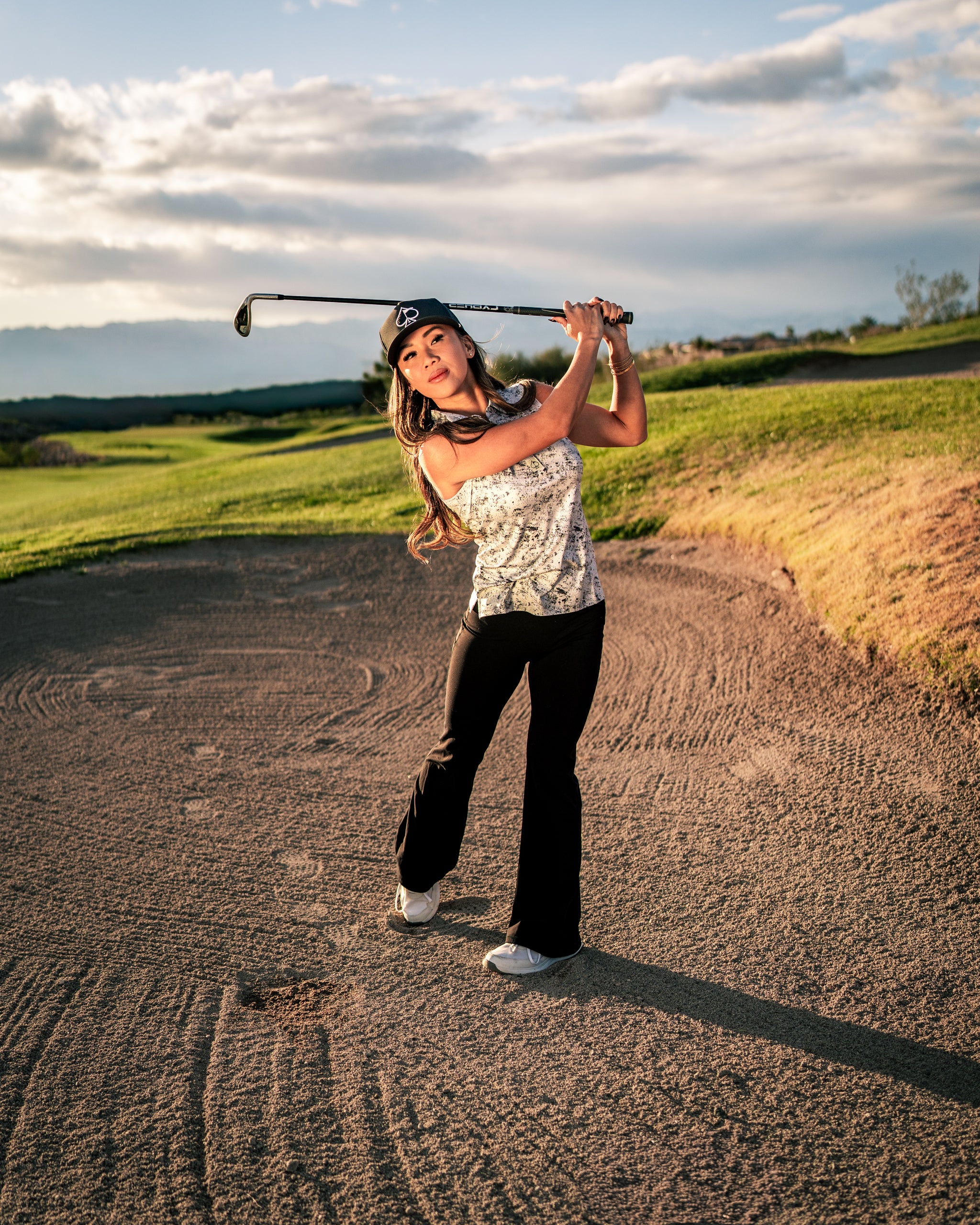 A woman swings a golf club on a course, wearing a stylish white noise top and black pants, showcasing bold golf fashion.