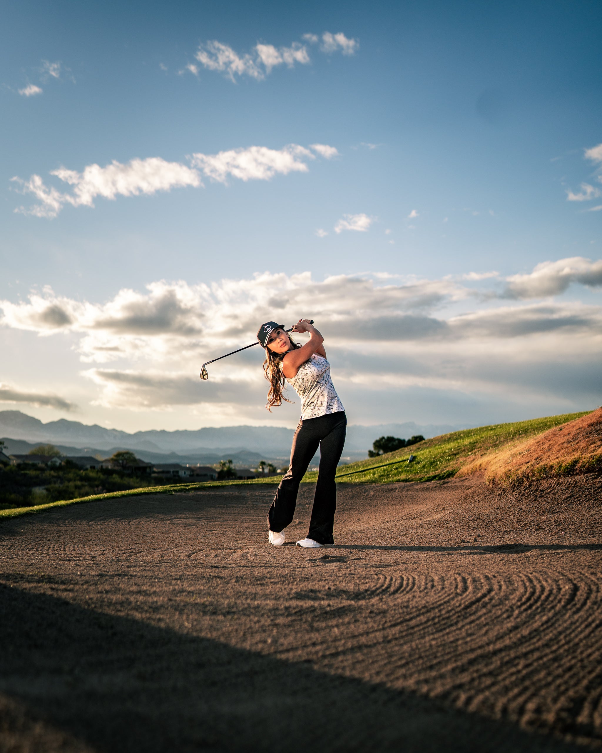 Woman in stylish golf apparel swings a club on a course with mountains in the background under a clear sky.