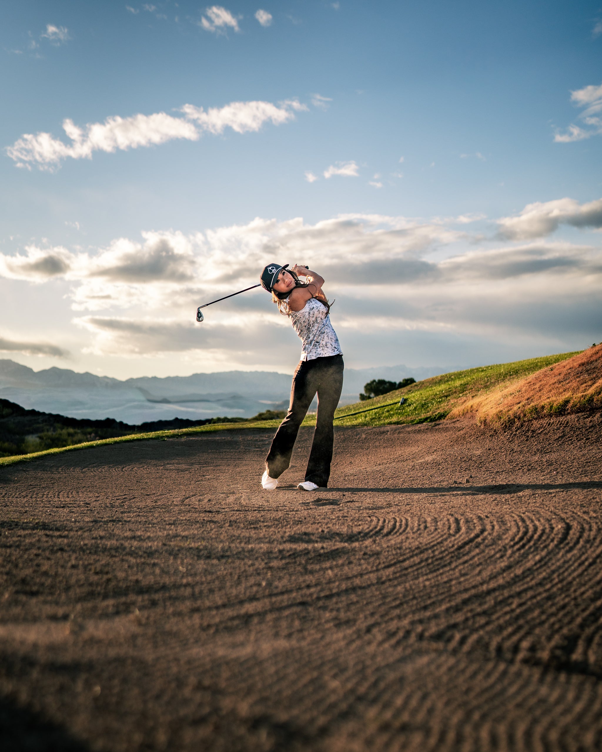 A woman swings a golf club on a sandy course, wearing a stylish white top and black pants, with mountains in the background.