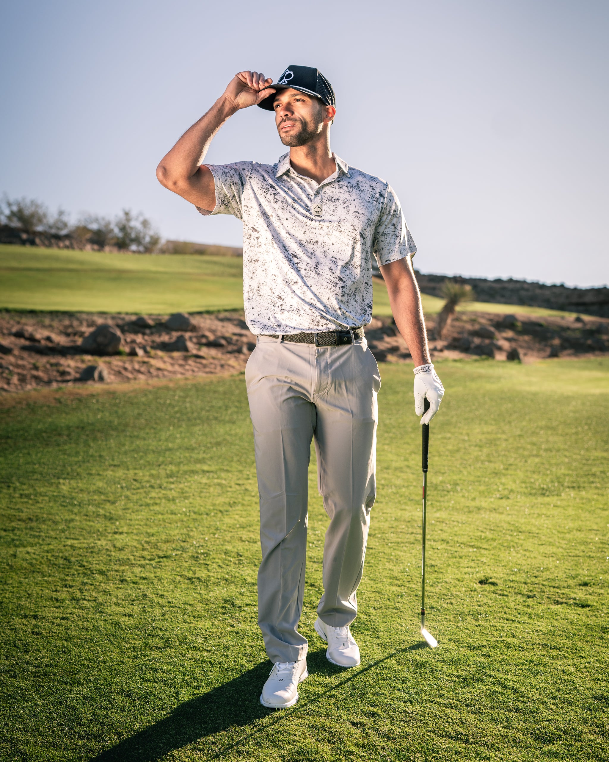 A golfer in a stylish white patterned shirt and light gray pants stands on the course, adjusting his cap.