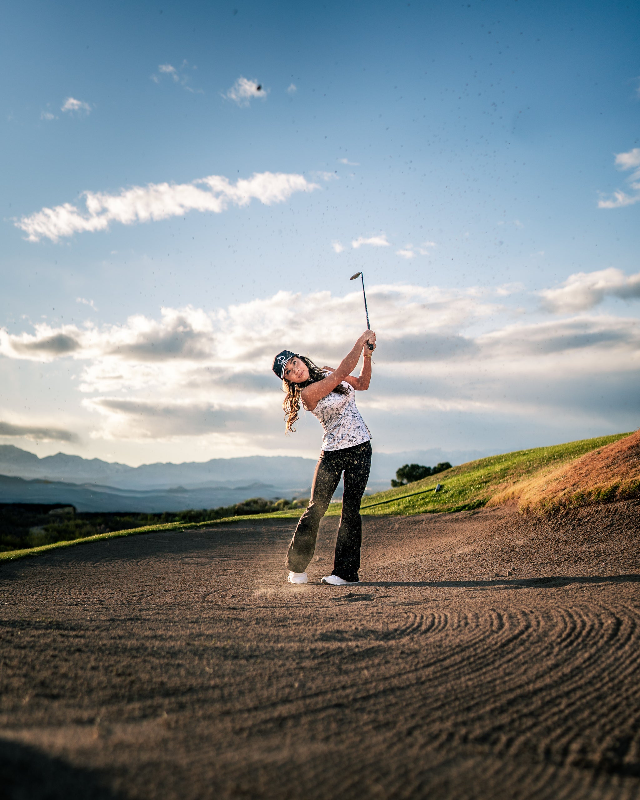 A woman swings a golf club on a sandy course, wearing a stylish white golf top and black pants against a scenic backdrop.