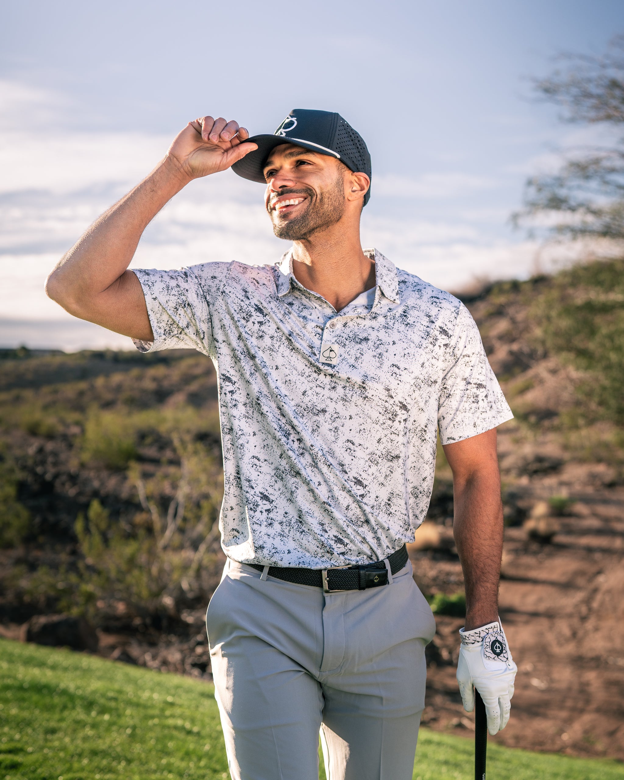 A golfer smiles while adjusting his cap, wearing a stylish white patterned polo shirt and light gray pants on a sunny course.
