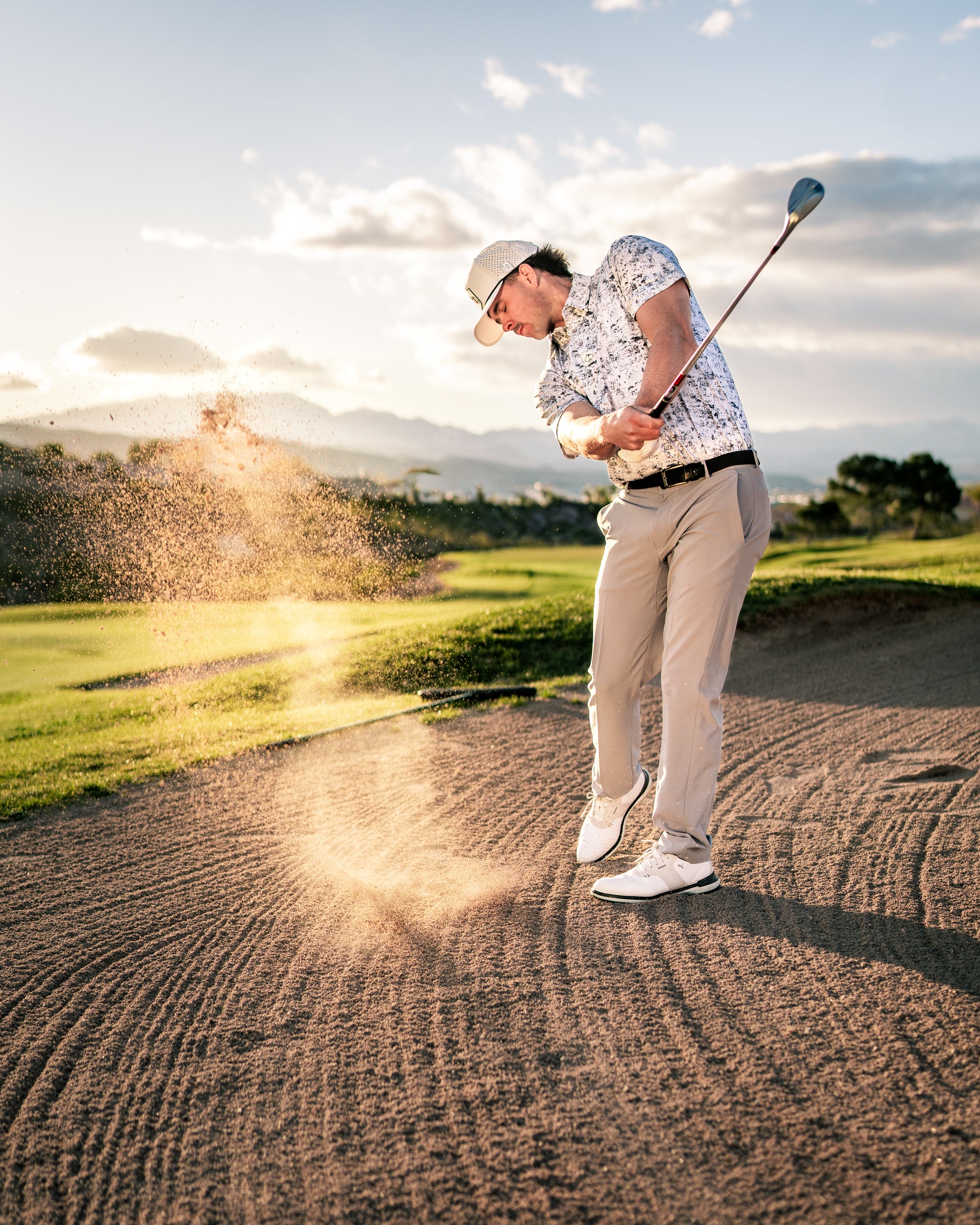 Golfer hitting a sand shot from a bunker, with sand flying in the air against a scenic backdrop of mountains and clouds.
