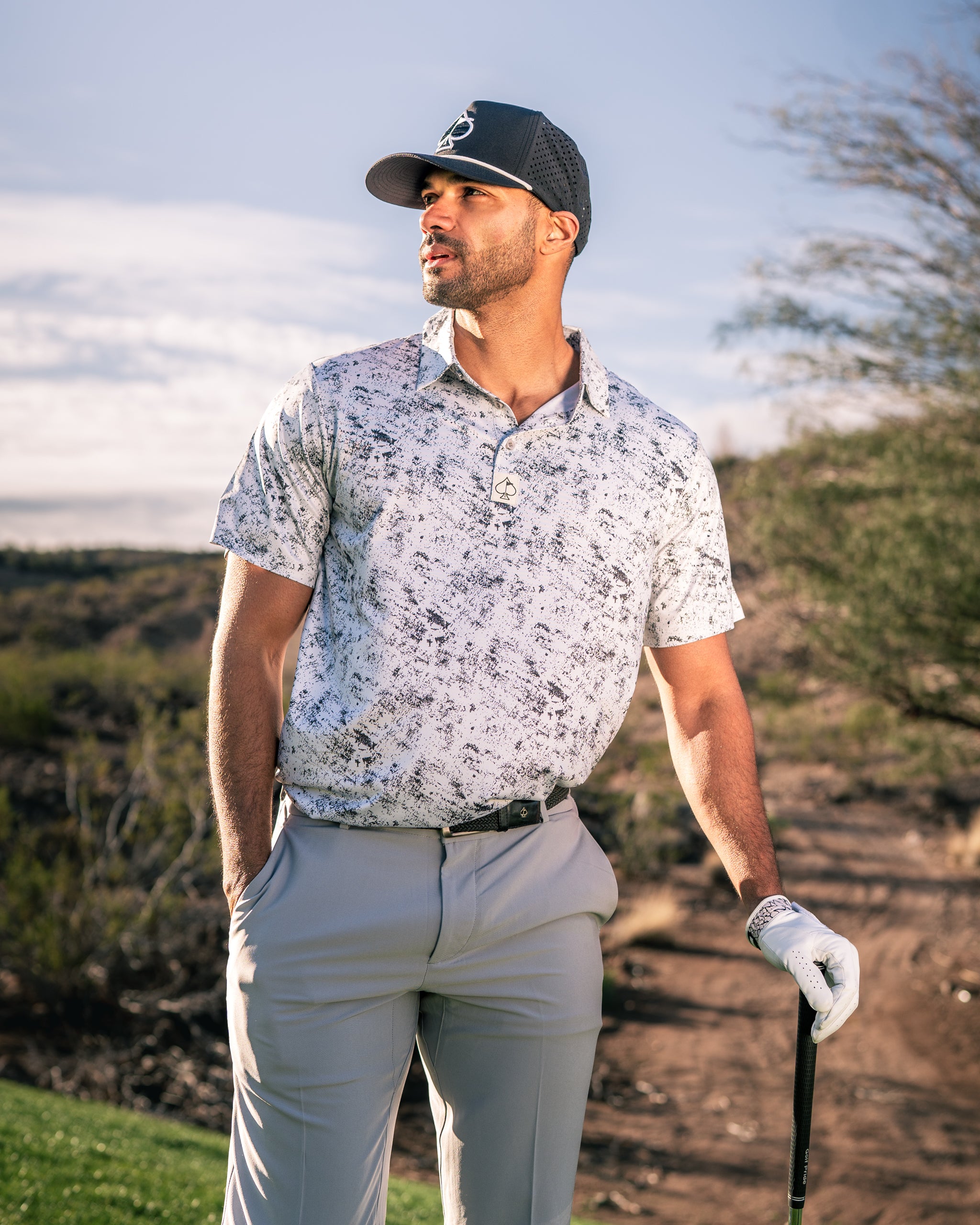 Stylish golfer wearing a white patterned polo shirt and gray pants, holding a golf club on a sunny course.