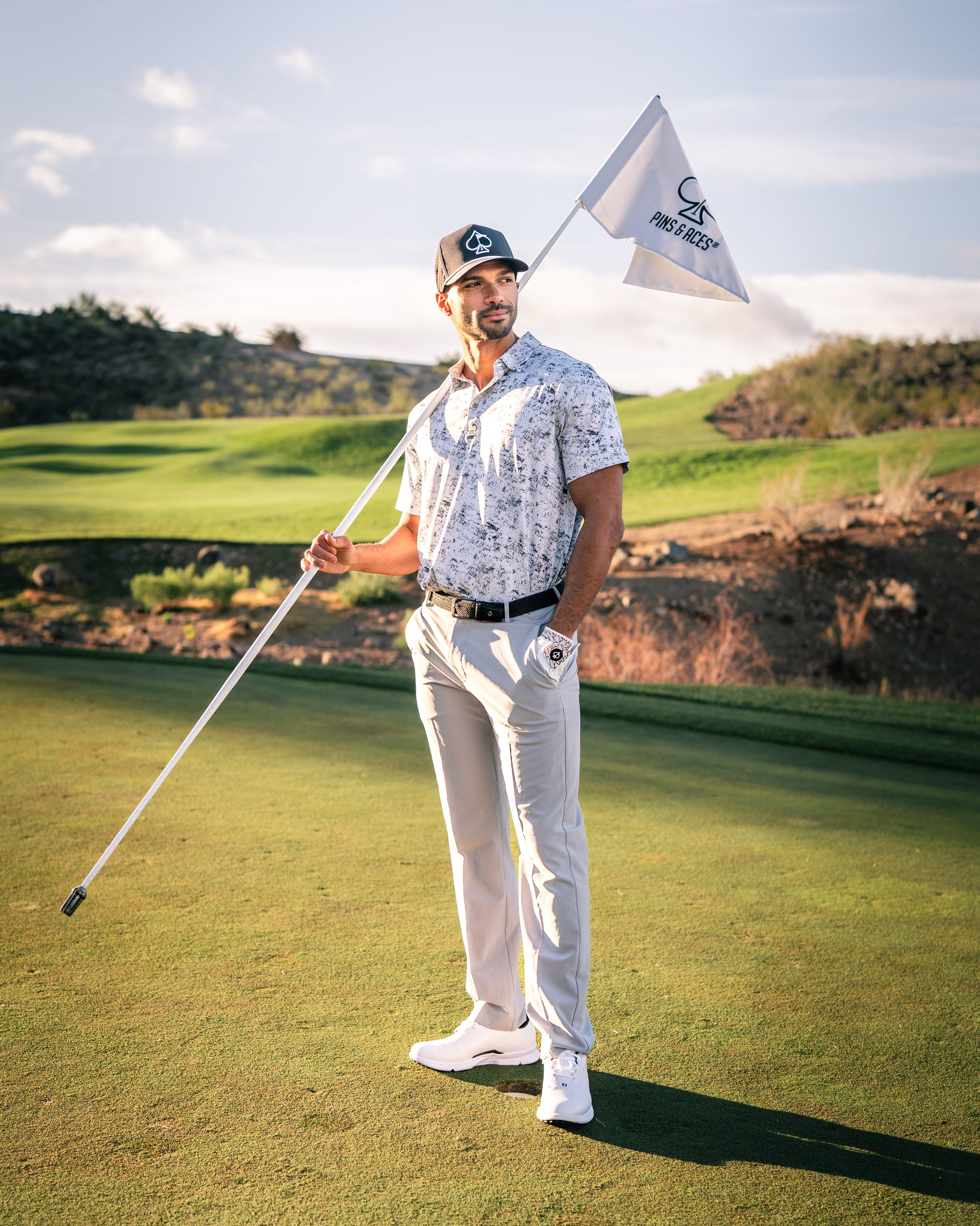 A golfer in a patterned polo shirt and light pants stands on the course, holding a flag with a logo, ready to play.
