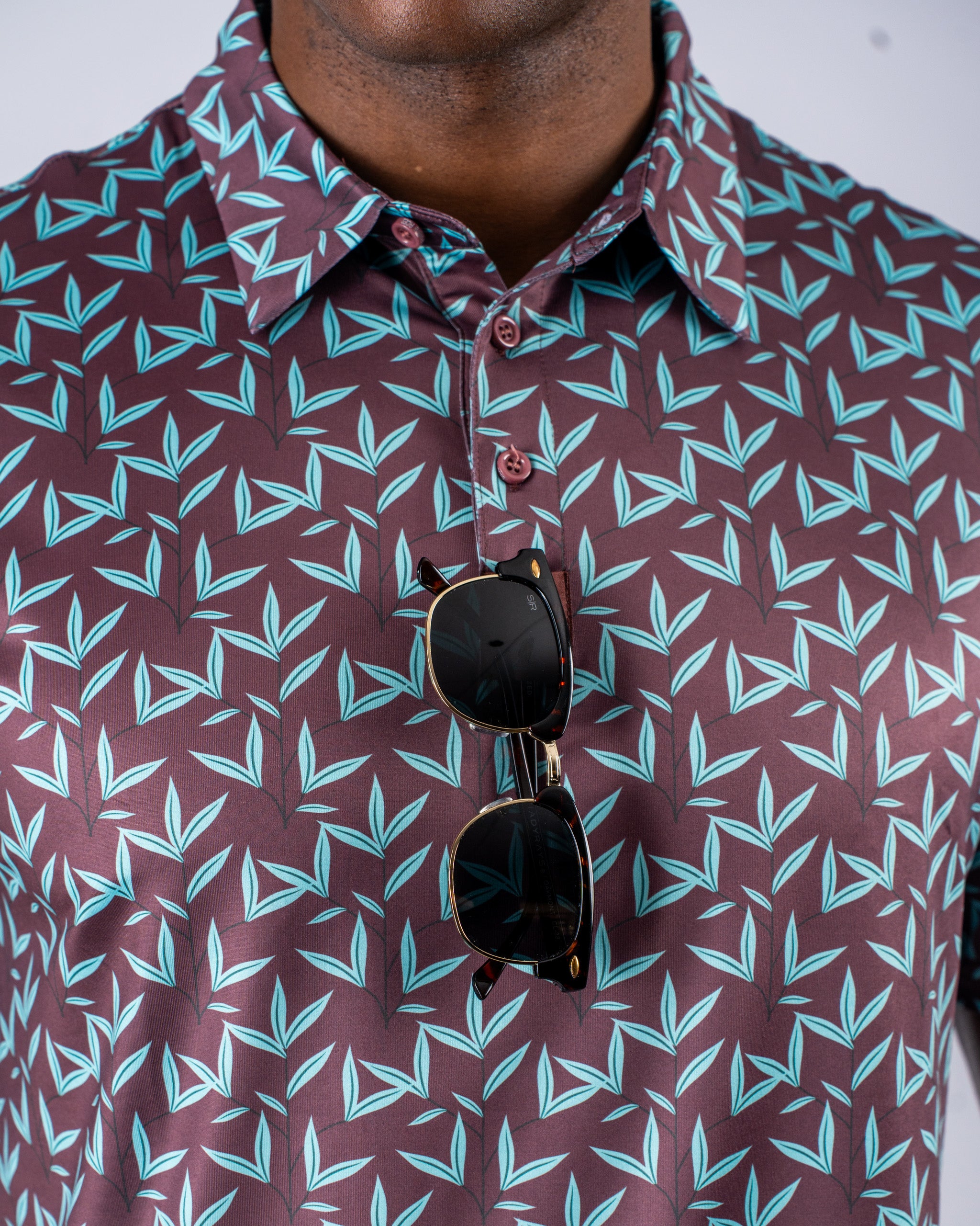 Bold golf shirt in cocoa brown with a teal leaf pattern, featuring a collar and sunglasses resting on the chest.