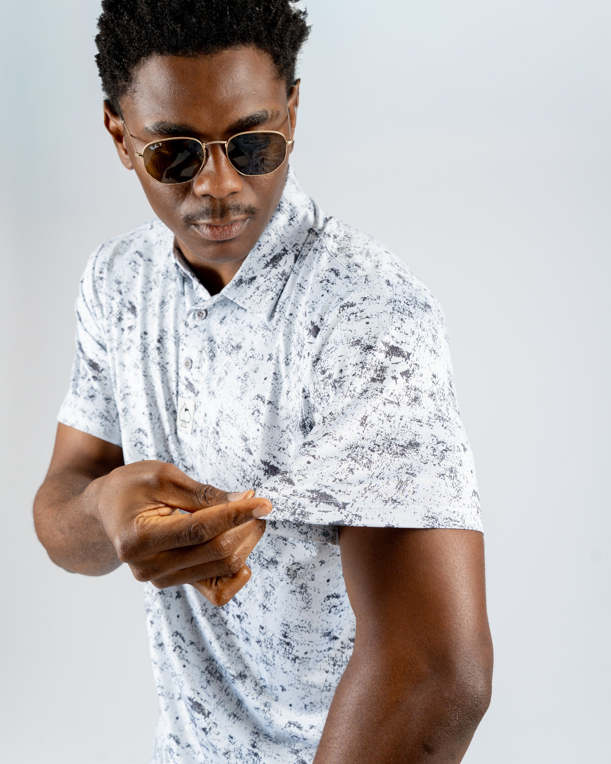 A man in sunglasses adjusts the sleeve of a white and gray patterned polo shirt, showcasing a stylish golf apparel look.
