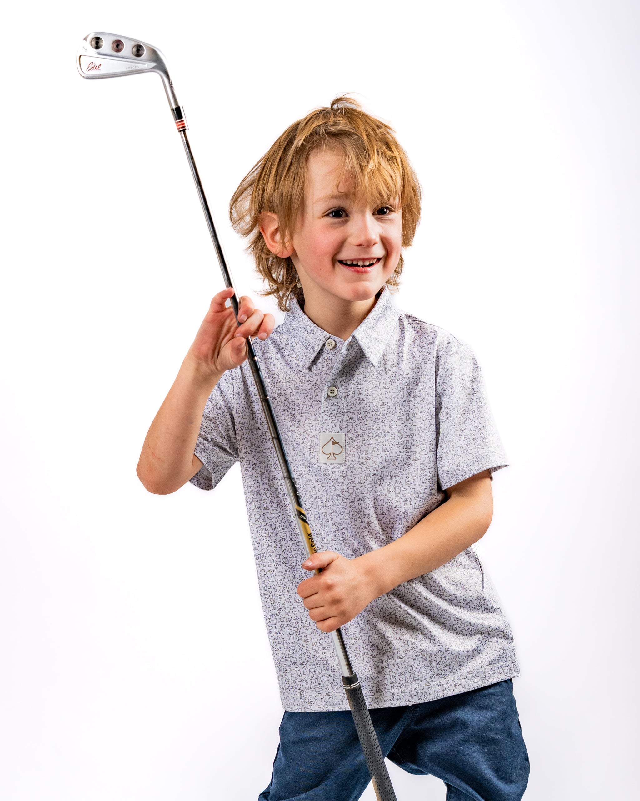 A young boy smiles while holding a golf club, wearing a stylish patterned polo shirt and casual pants.