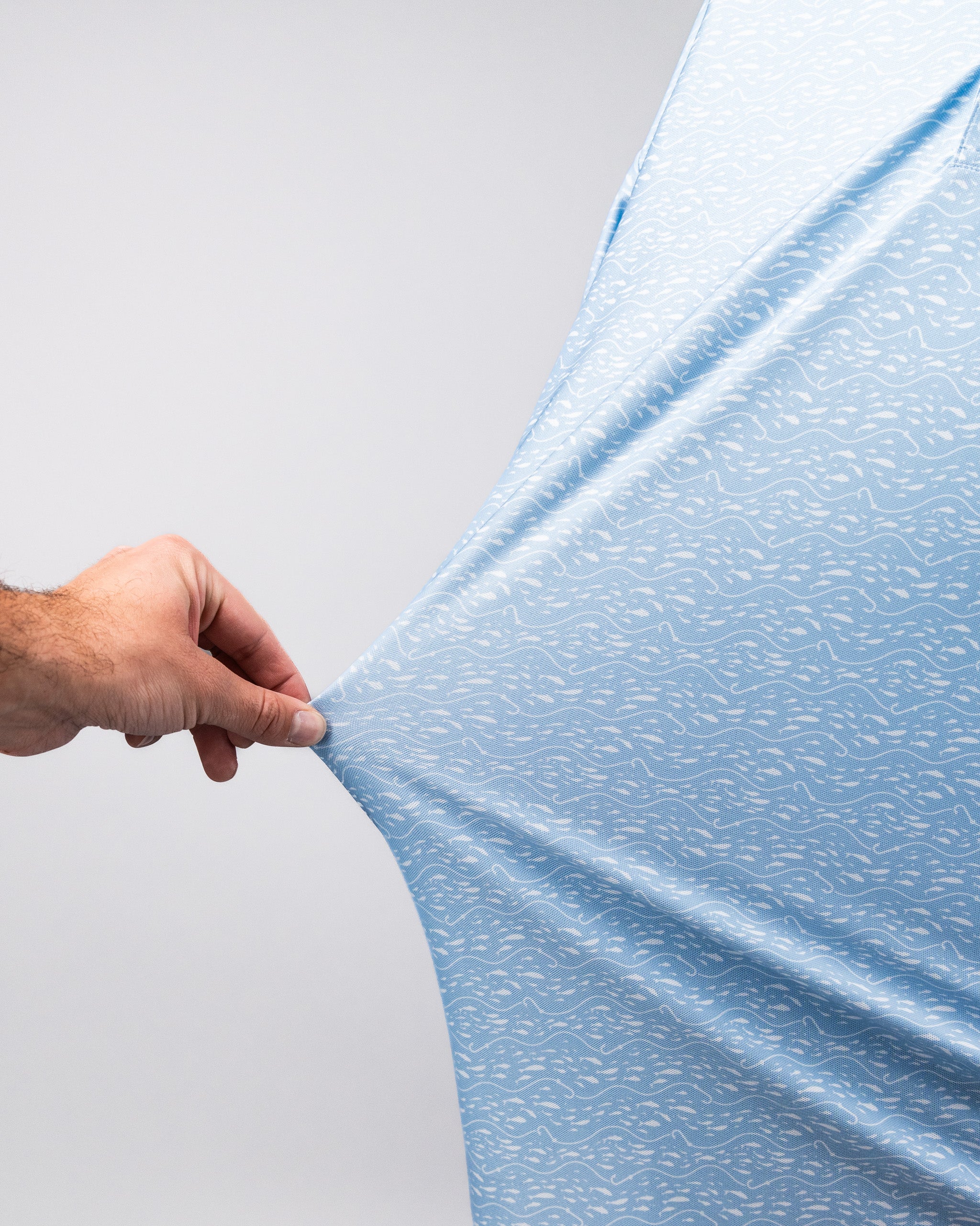 Close-up of a hand pulling a light blue Player Preferred™ Polo - Tidal Flow fabric, showcasing its textured, water-inspired pattern.