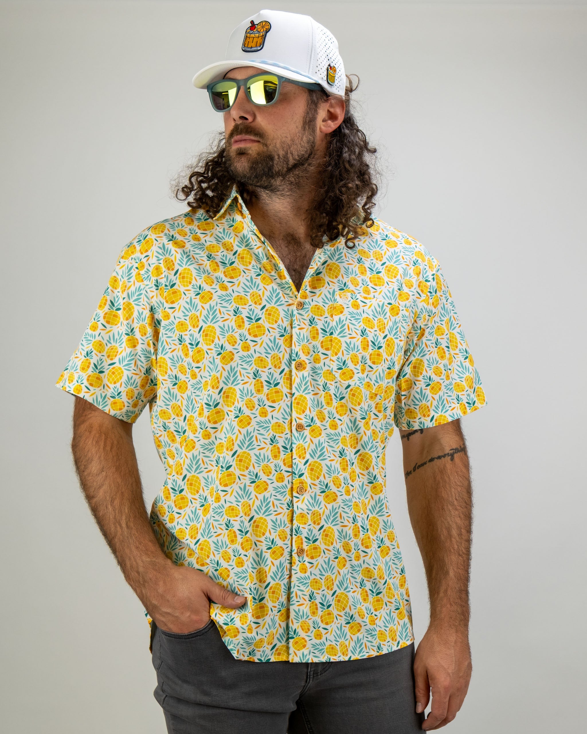 Bold short-sleeve shirt featuring a vibrant pineapple pattern, paired with a white cap and sunglasses for a stylish golf look.