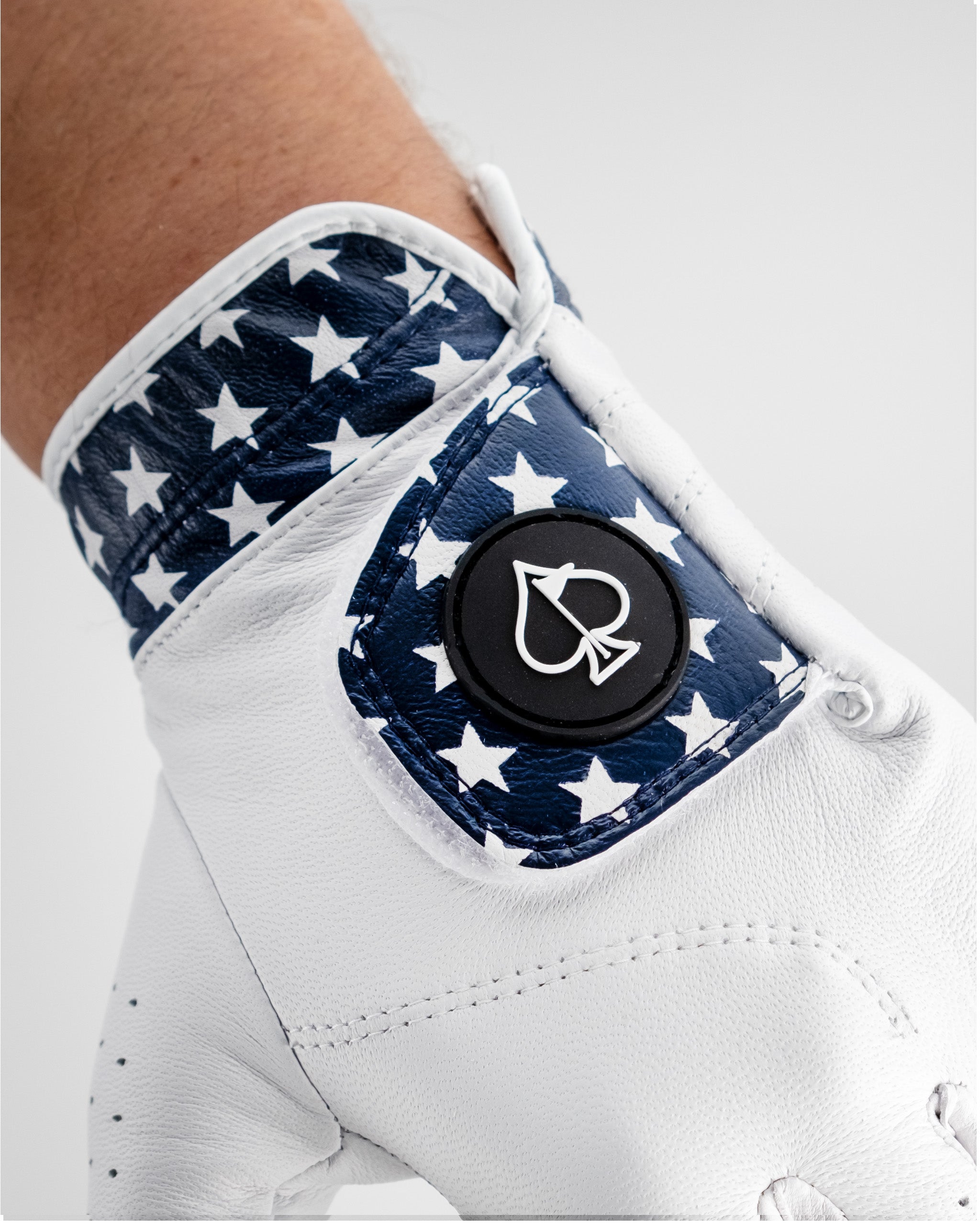Close-up of a white golf glove with a navy blue star pattern and a black circular logo patch on the wrist.