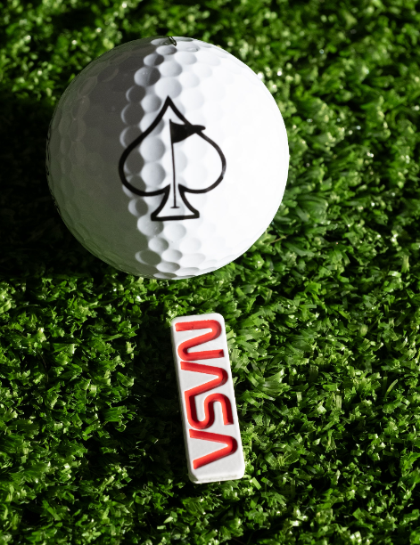 NASA "Worm" ball marker positioned next to a golf ball featuring a spade logo, set against a green turf background.