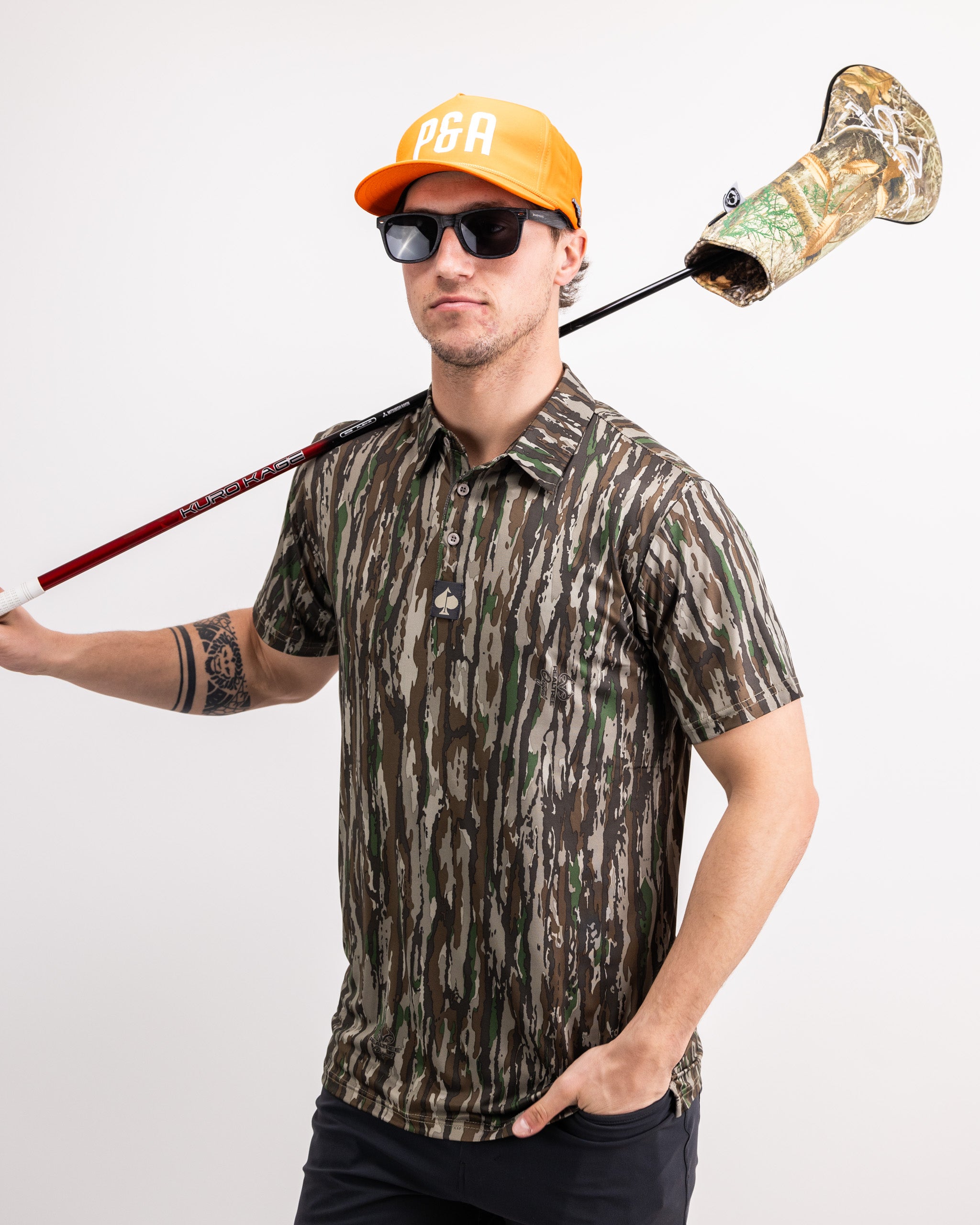 Man wearing a Realtree Original Polo, holding a golf club with a camo headcover, sporting sunglasses and a bright cap.