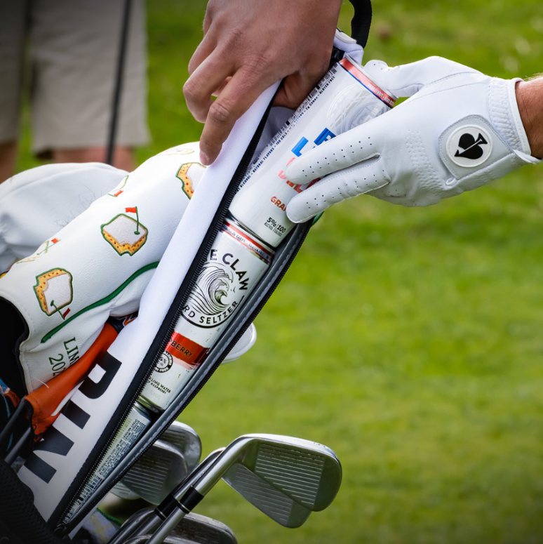 A golfer's hand reaches into a golf bag to retrieve a can from a stylish seltzer sleeve, showcasing bold patterns.