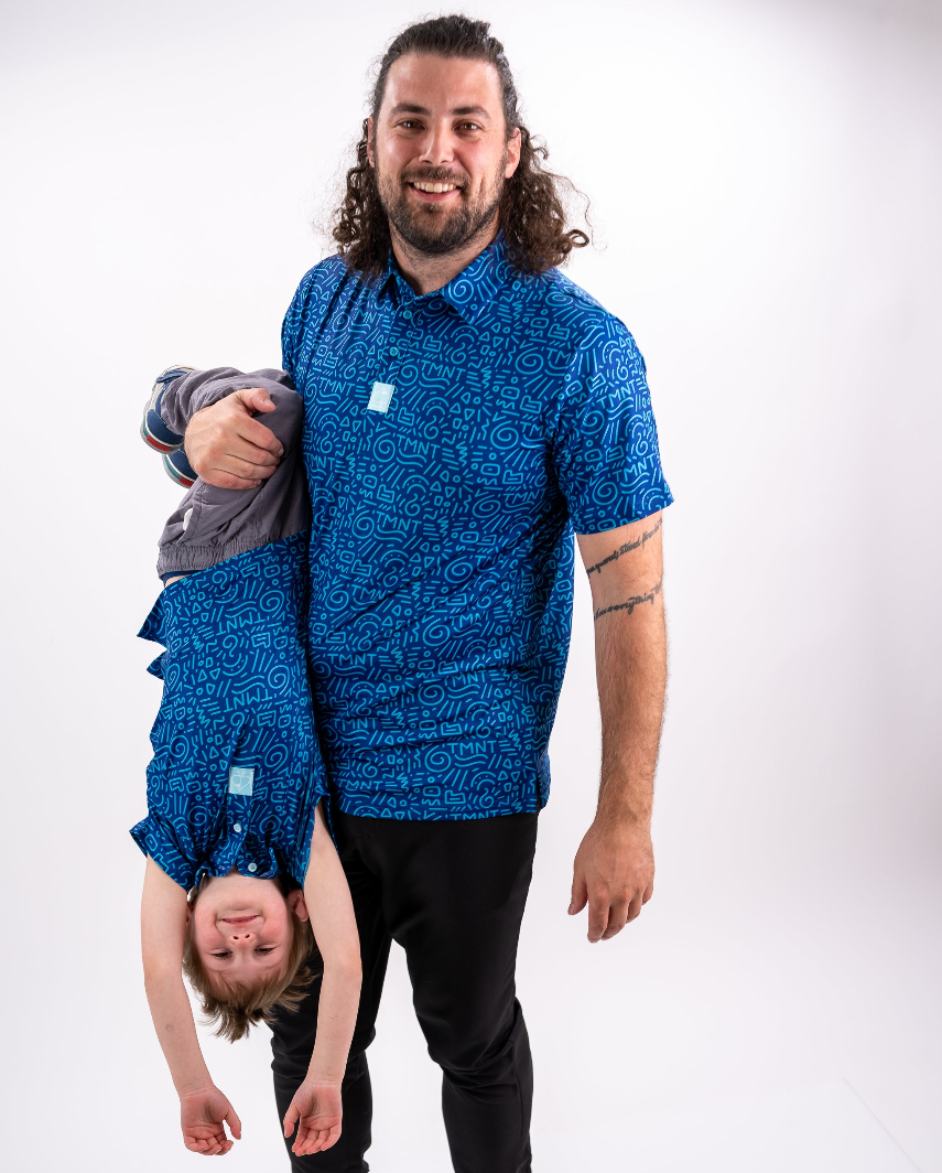 A man holds a child upside down, both wearing matching blue TMNT doodle-patterned shirts, showcasing playful family fun.