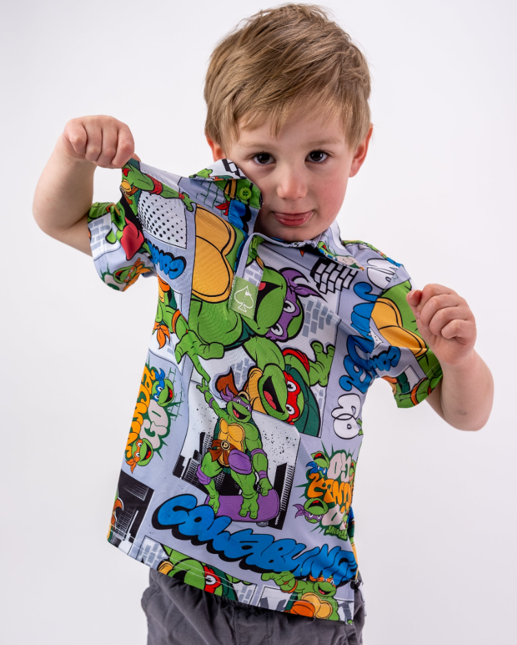 A young child shows off a colorful TMNT comic strip shirt, featuring vibrant graphics of the Teenage Mutant Ninja Turtles.