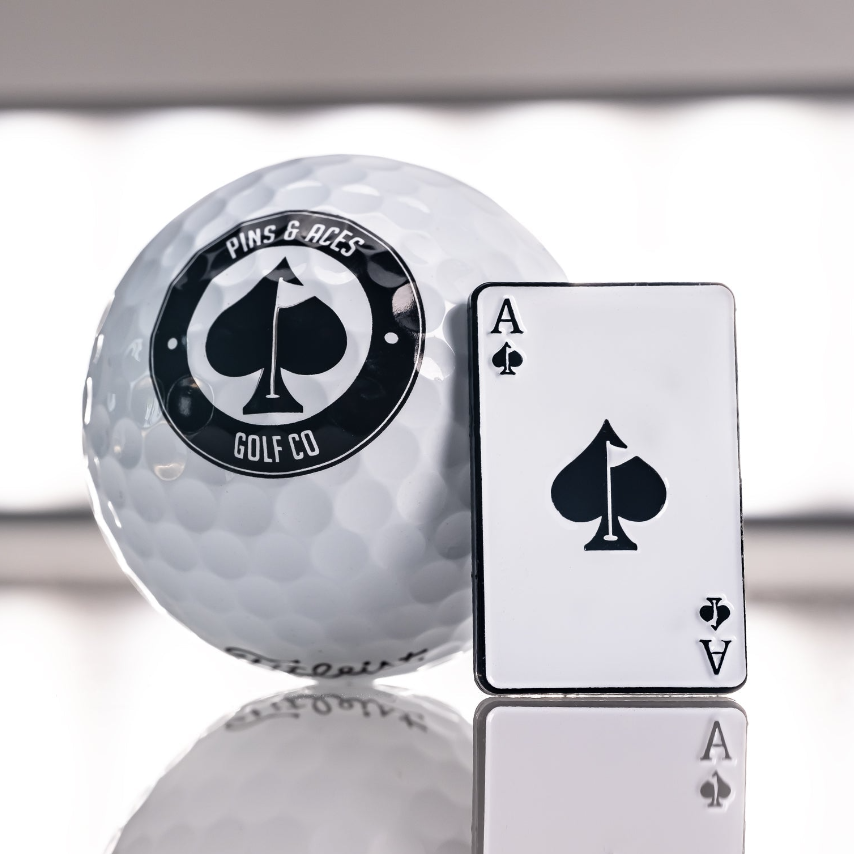 Golf ball with "Pins & Aces Golf Co" logo and an Ace of Spades playing card, showcasing bold, unique golf accessories.