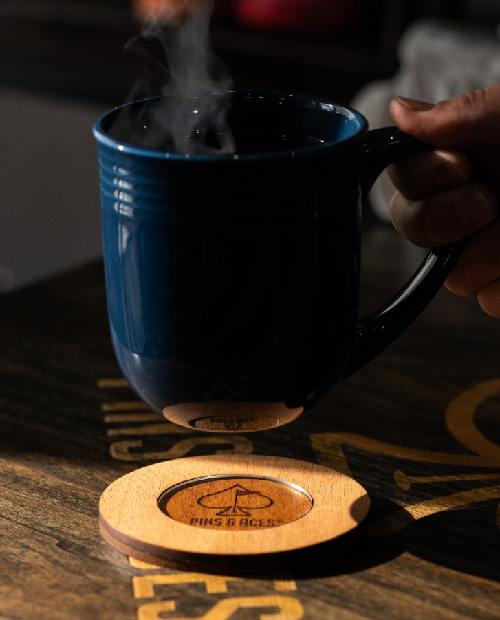 Wooden coaster from Pins & Aces with logo, holding a steaming mug above it on a wooden surface.