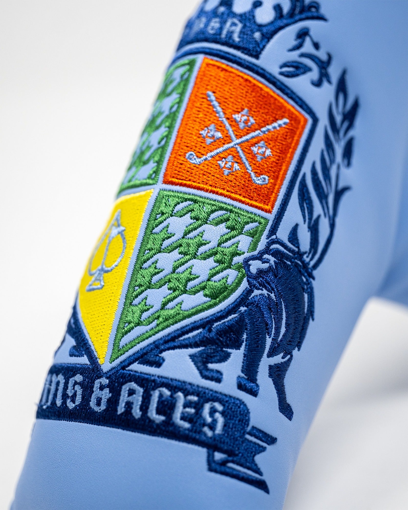 Bold blue blade putter cover featuring an intricately embroidered coat of arms design with golf motifs and vibrant colors.