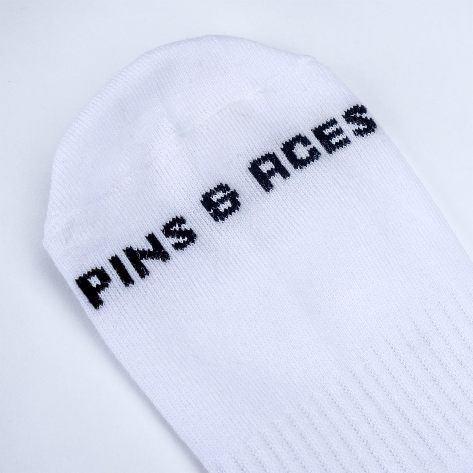 White ankle socks featuring the brand name "Pins & Aces" in bold black lettering. Ideal for stylish golf enthusiasts.