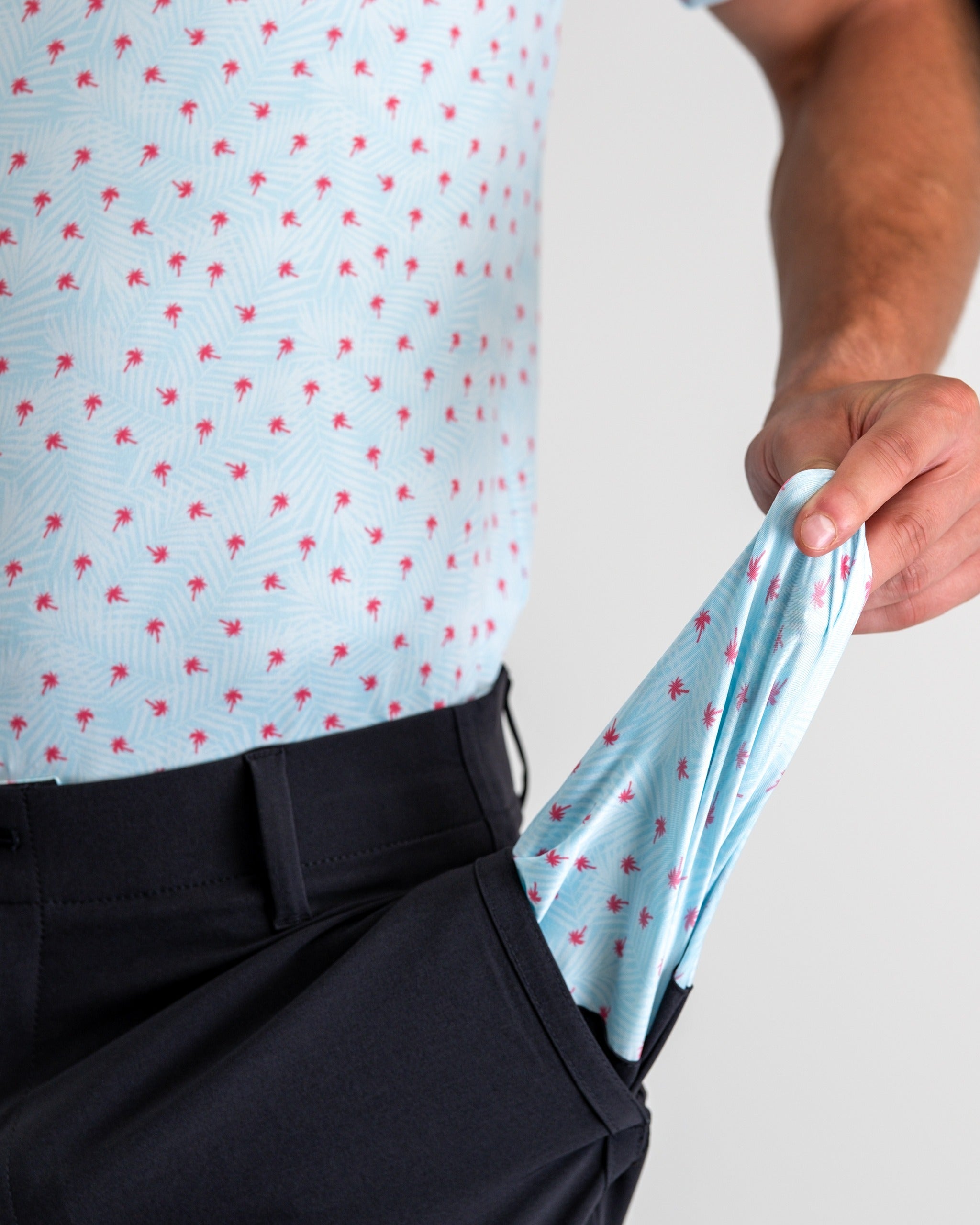 Men's black performance shorts with a hand pulling out a light blue shirt featuring a palm tree pattern. Ideal for golf.